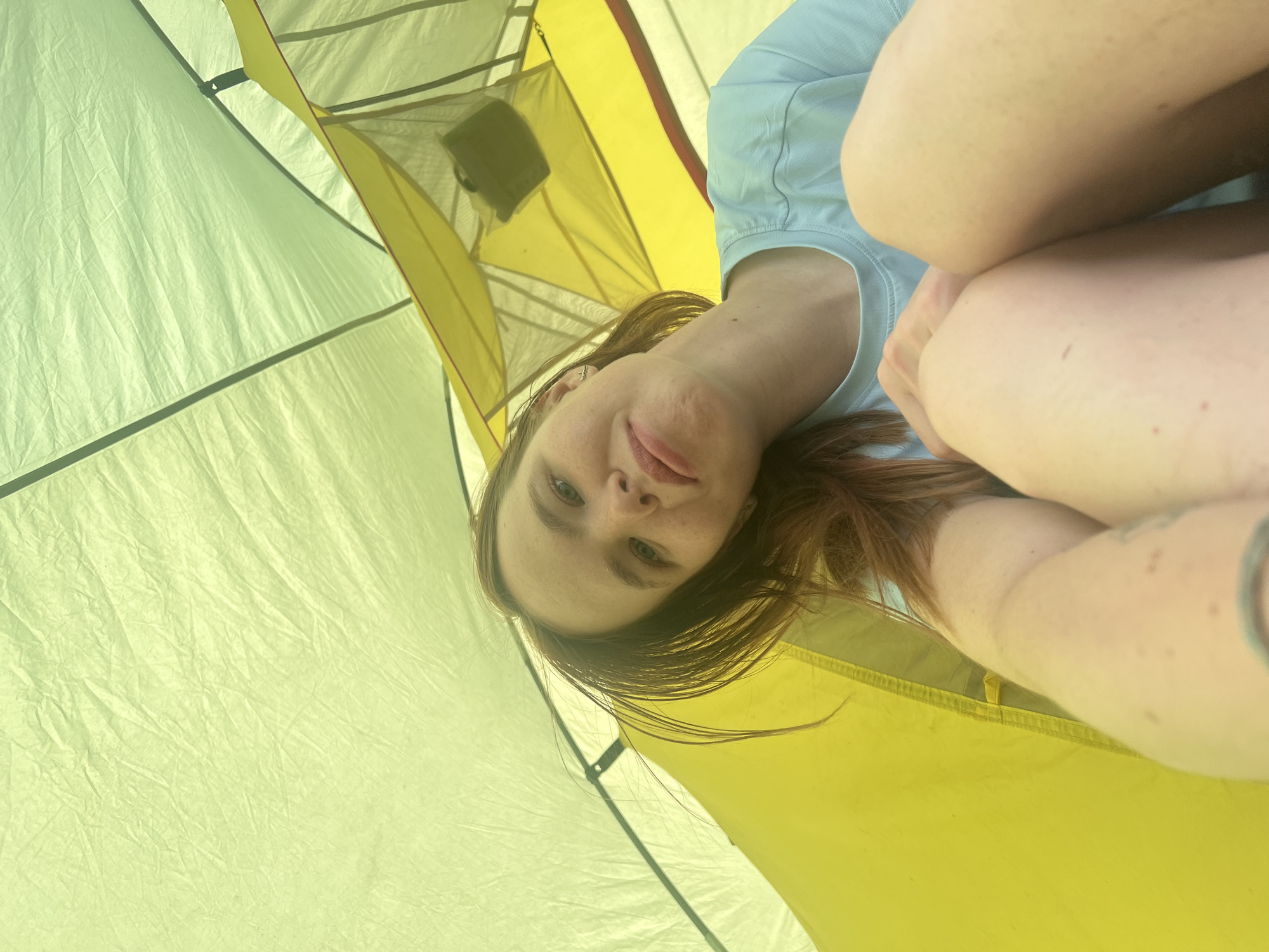 A woman with red hair and fair skin lies inside a yellow and green tent. She is wearing a light blue shirt and has a tattoo on her arm. The tent is located in an unknown location.