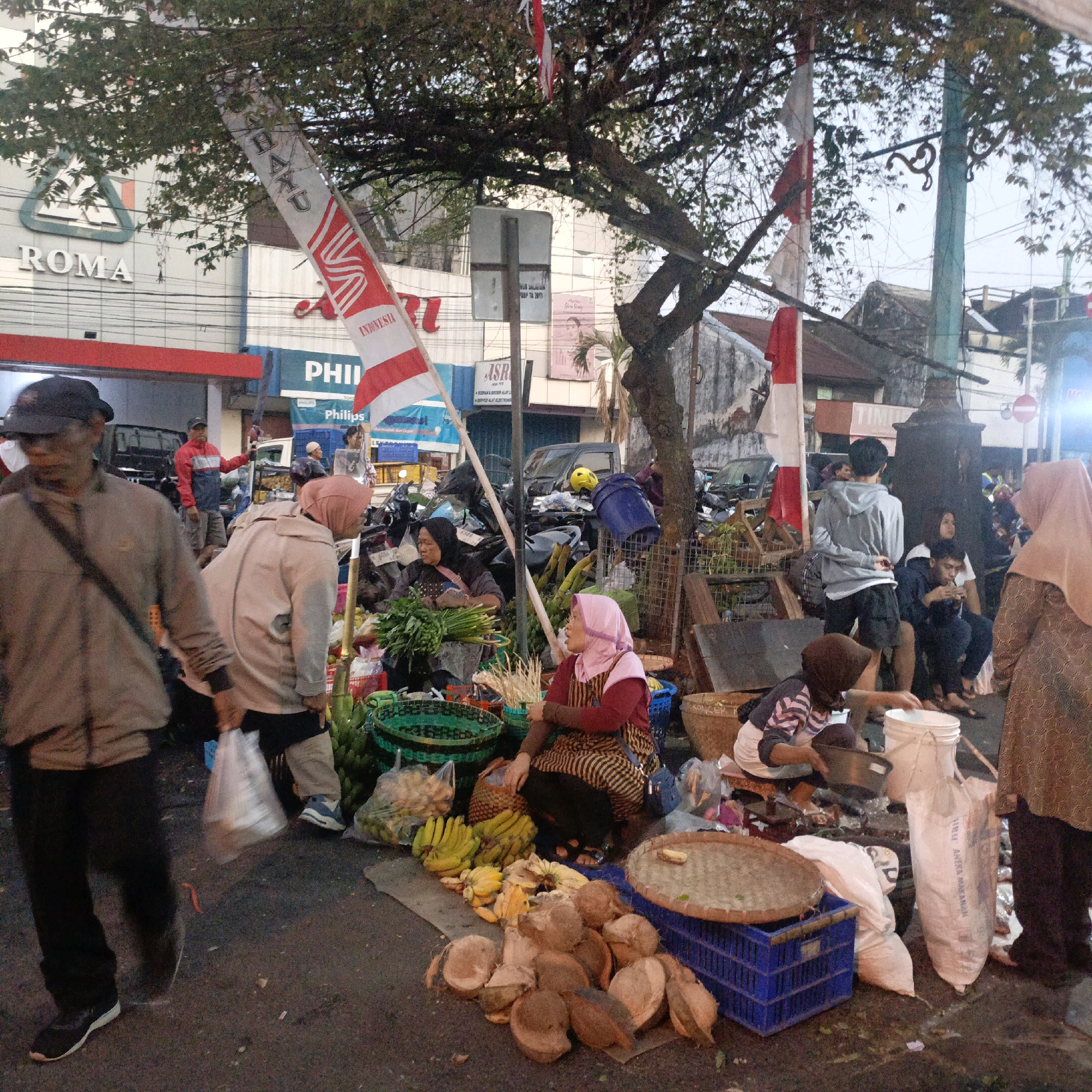 The image depicts a street scene in Salatiga, Indonesia, likely a market or roadside stall area. In the foreground, several individuals are visible. A man wearing a dark cap and a gray jacket is walking towards the left of the frame. Behind him, a woman in a light-colored hijab and jacket appears to be interacting with merchandise, possibly buying or selling. Further into the scene, a woman in a pink headscarf and a striped dress is seated, attending to a display of fruits and coconuts.
The environment is cluttered with various items. Piles of fruits, including bananas, are arranged on the ground and in baskets. Coconut shells are scattered around. Several bags, some filled with produce and others appearing to be sacks, are present. In the background, a building with the word "ROMA" prominently displayed on its facade suggests a commercial establishment. Other buildings and a street scene with vehicles and motorcycles are also visible. Indonesian flags are hung, adding a nationalistic element to the setting. The lighting suggests it might be late afternoon or early evening. The overall impression is a bustling local market environment.