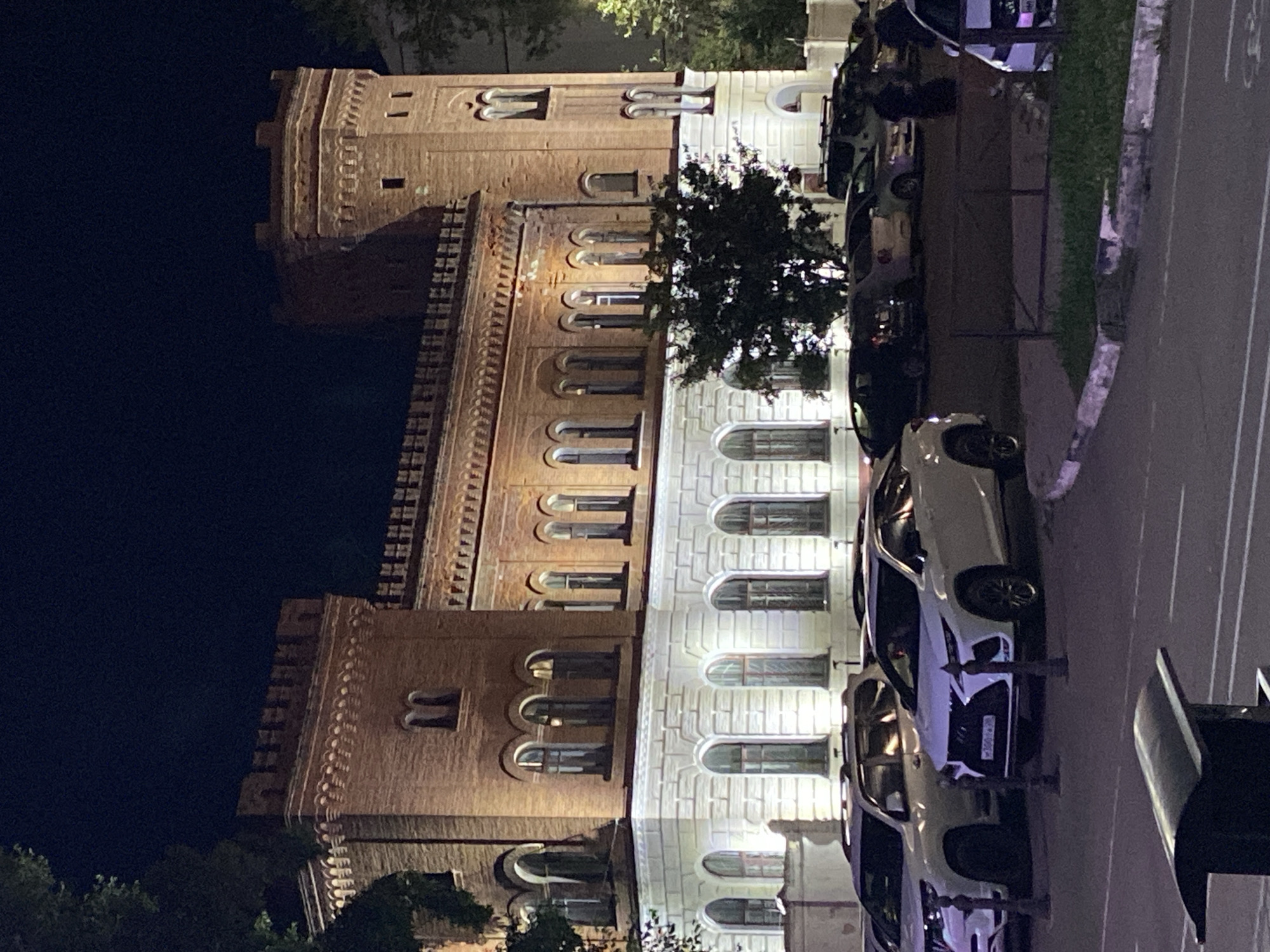 The image shows a brick building with crenellated towers, possibly an old fortress or administrative building, illuminated at night. The building has arched windows on both the upper brick section and the lower white stone section. In front of the building, several cars are parked, including white SUVs and sedans. A dark-colored tree is visible in front of the building, and other trees and foliage are present on the right side of the image. The scene appears to be a street or parking area in an urban environment. This building is located at **25 Lenina Street, Blagoveshchensk, Russia**, and is known as the **Blagoveshchensk City Duma**.