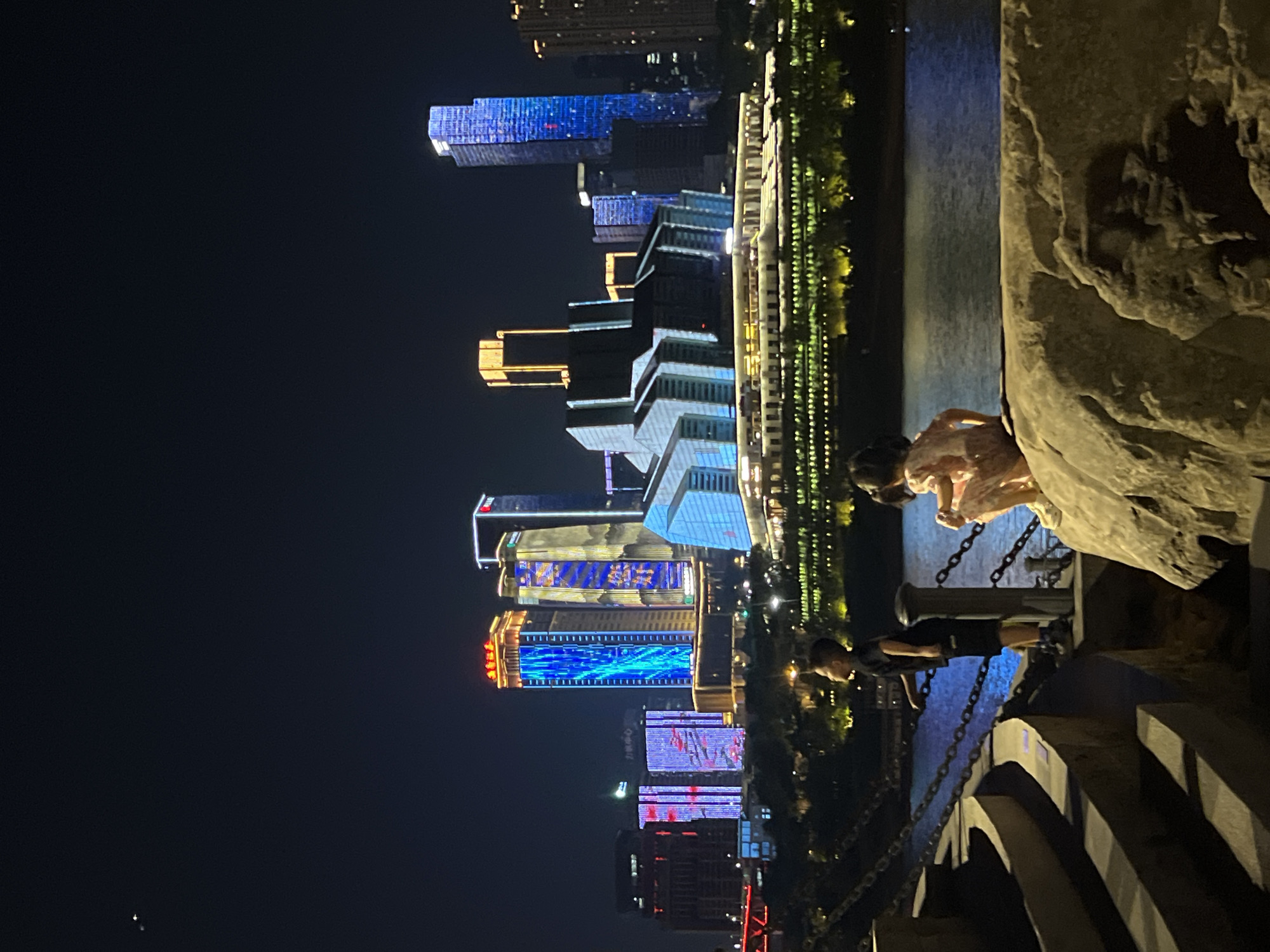 This is a nighttime photograph taken from a waterfront area in Yuzhong District, China. In the foreground, a boy stands facing right, looking towards the water, with his back to the camera. He is wearing a black t-shirt and black shorts. To his right, a young girl is seated on a large rock, facing away from the camera and looking towards the city skyline. She is wearing a pink dress. Behind them is a body of water, likely a river, reflecting the city lights.
Across the water, a modern cityscape is illuminated by numerous lights. Prominent are several tall buildings, some with dynamic LED displays. One building in the center has a striking facade of blue and white lights, while others feature golden and red illumination. A large, angular building with a bright, illuminated roof structure is situated in front of some of the taller buildings. The cityscape is bordered by a line of trees and lights along the riverbank, creating a festive and vibrant atmosphere. The sky is dark, with two faint celestial objects visible in the upper left corner. The scene depicts a moment of quiet observation by the children amidst a bustling urban backdrop.