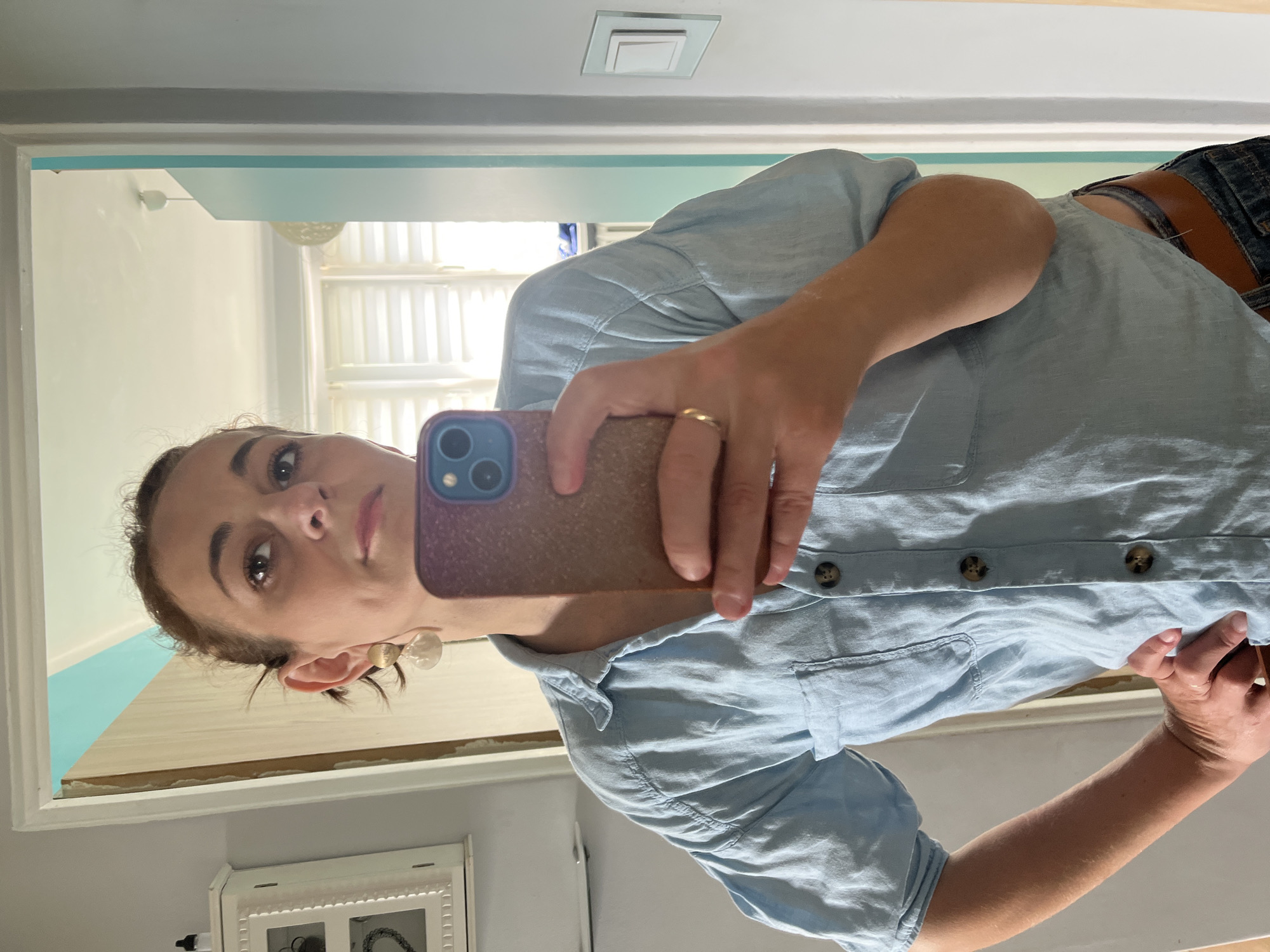 A woman takes a selfie in a mirror, holding her phone in her right hand. She is wearing a light blue button-down shirt and denim shorts with a brown belt. The reflection shows her face, with dark hair pulled back and large hoop earrings. She is wearing a ring on her left ring finger. The background of the mirror's reflection shows a room with light blue walls and white blinds on a window. A light fixture is visible on the ceiling. The mirror itself is framed in white. The overall impression is a casual, everyday moment captured at home.