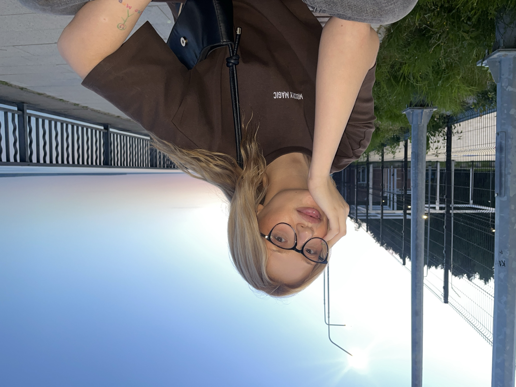 A young, fair-skinned woman with blonde hair and black-framed glasses is pictured outdoors in Chaikovsky, Russia. She is wearing a brown t-shirt with the words "MOSTLY MAGIC" printed on it and a black shoulder bag. The backdrop includes a sky gradient from blue to white due to sunlight, a railing overlooking what appears to be a body of water, a fence with metal poles, and some greenery. The image is oriented upside down.