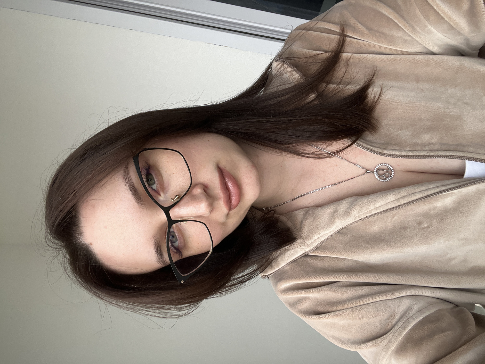 A close-up portrait features a young woman, likely taken indoors in Tyumen, Russia. She has long, dark brown hair that frames her face, and wears stylish black-framed glasses. Her light-colored eyes, appearing green or hazel, have a subtle touch of pinkish eyeliner on the lower lash line. Her lips have a natural, glossy finish. She maintains a calm, neutral expression with a hint of a soft smile.
She is dressed in a soft, beige or light brown velour or velvet zippered top or jacket. A silver chain necklace adorns her neck, featuring a circular pendant with the 'Leo' zodiac symbol at its center, surrounded by small clear stones. The background consists of a plain, light-colored wall, with a thin white window frame visible at the upper edge of the image, suggesting an indoor setting. The lighting is soft and even, indicating diffused natural light or gentle indoor illumination. The photograph captures her looking towards the camera, suggesting a selfie or a candid portrait.