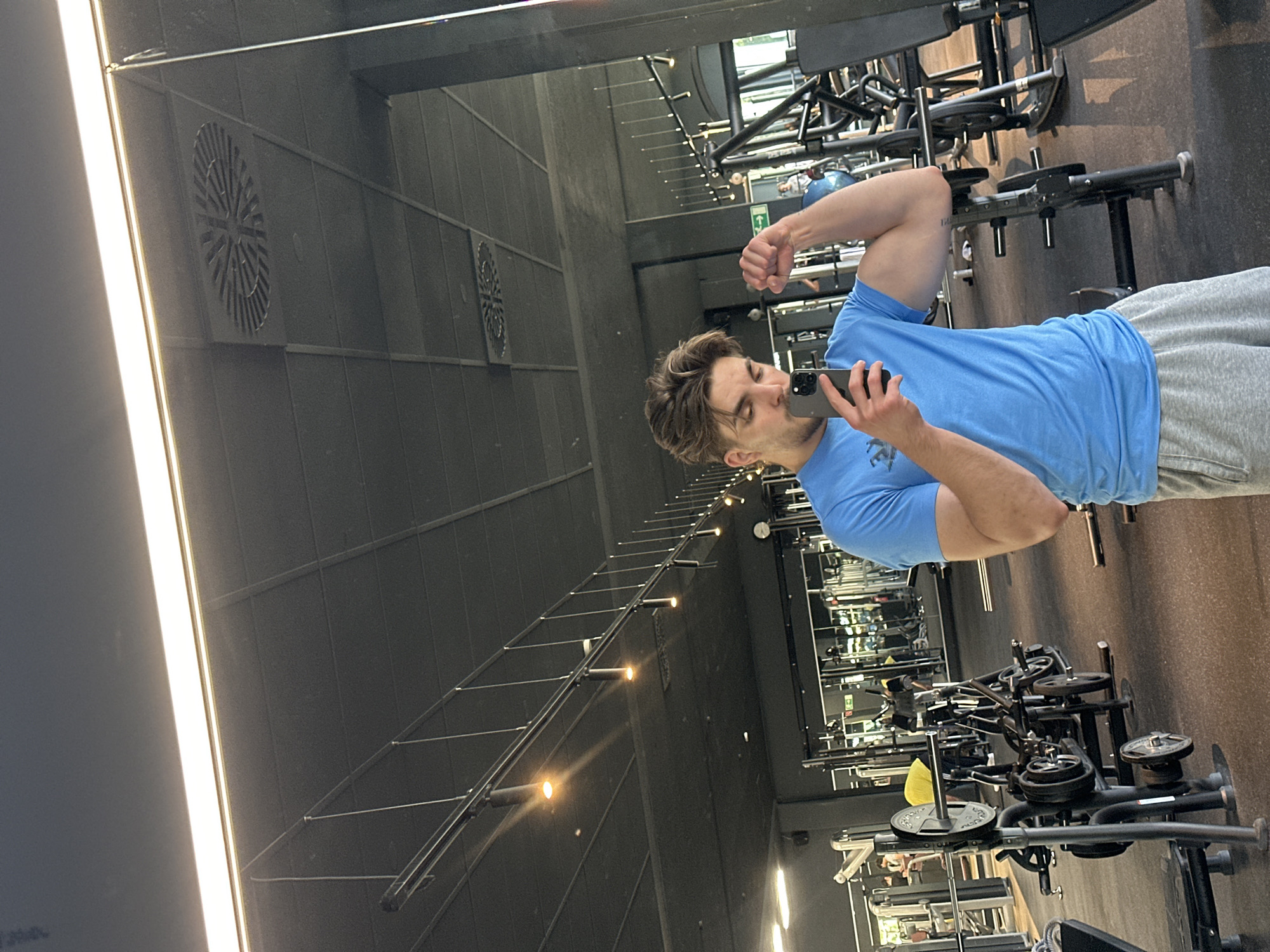 A fair-skinned man with brown hair is taking a selfie in a mirror at a gym. He is wearing a light blue t-shirt and gray sweatpants. He is holding a smartphone in his left hand and flexing his right bicep. Gym equipment is visible in the foreground and background, including weight racks, benches, and machines. The gym has black walls with vents, and there are track lighting fixtures along the ceiling. The photograph was taken in Jestetten, Germany.