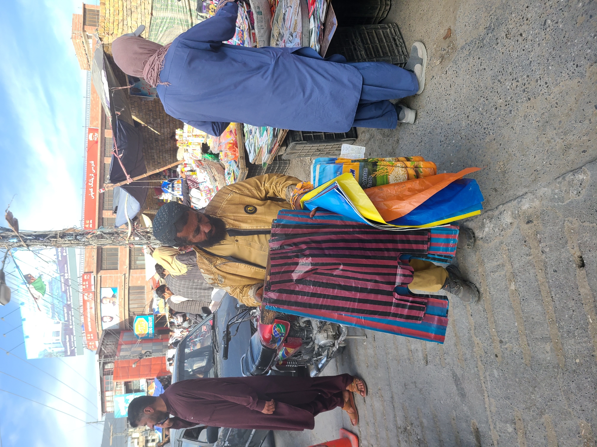 The scene takes place in an outdoor market area of Quetta City Tehsil, Pakistan, on a paved road or street. A man with a beard, wearing a tan jacket and holding bags in front of him, appears to be selling goods. There is a blue motorcycle visible behind the man, and another man walks nearby in a maroon robe and sandals. Another person is standing to the left, wearing a light-colored robe. Additionally, there are a couple of vendors displaying products under makeshift stalls, with colorful kites and other merchandise visible. Buildings and billboards are in the background of the image.