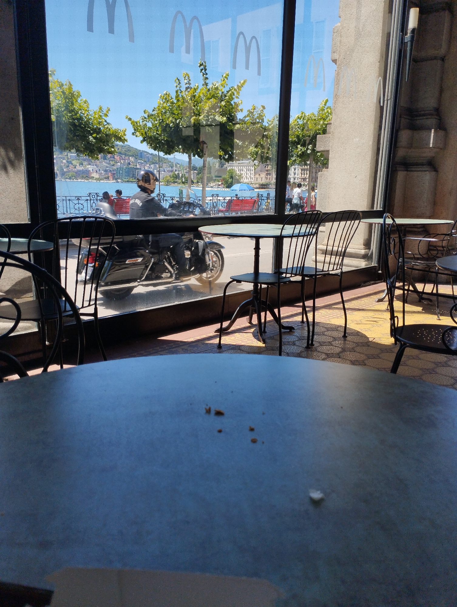 The image shows a reflection of a street scene through a large window, likely from inside a McDonald's restaurant in Lugano, Switzerland. The reflection displays a bright blue sky, lush green trees, and a distant cityscape with buildings situated along a body of water, presumably Lake Lugano. A man wearing a helmet is riding a black motorcycle in the foreground of the reflection, with his back to the viewer. In front of him, two individuals are seated on a red bench, also reflected. Several empty tables and chairs, characteristic of outdoor seating at a cafe or restaurant, are visible in the foreground. The floor inside the establishment appears to be tiled with a patterned design. The iconic McDonald's logo is subtly visible as a reflection on the glass.