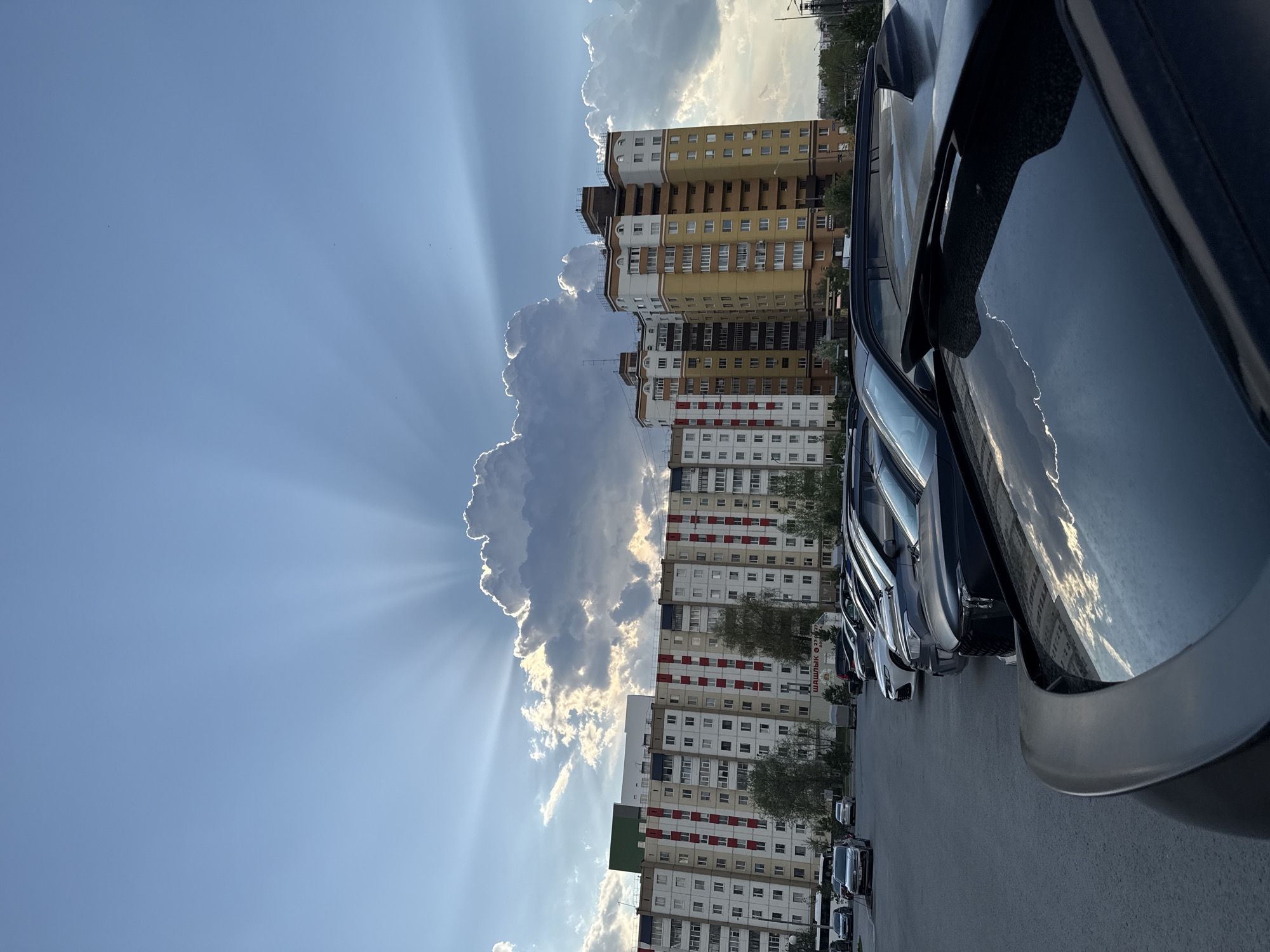 The image shows a row of parked cars along a street with several tall apartment buildings in Tobolsk, Russia. The buildings are predominantly white with some having red and tan accents. A large cumulus cloud is visible in the sky, with sun rays streaming from behind it. The sky is blue. The scene appears to be taken during the day.
