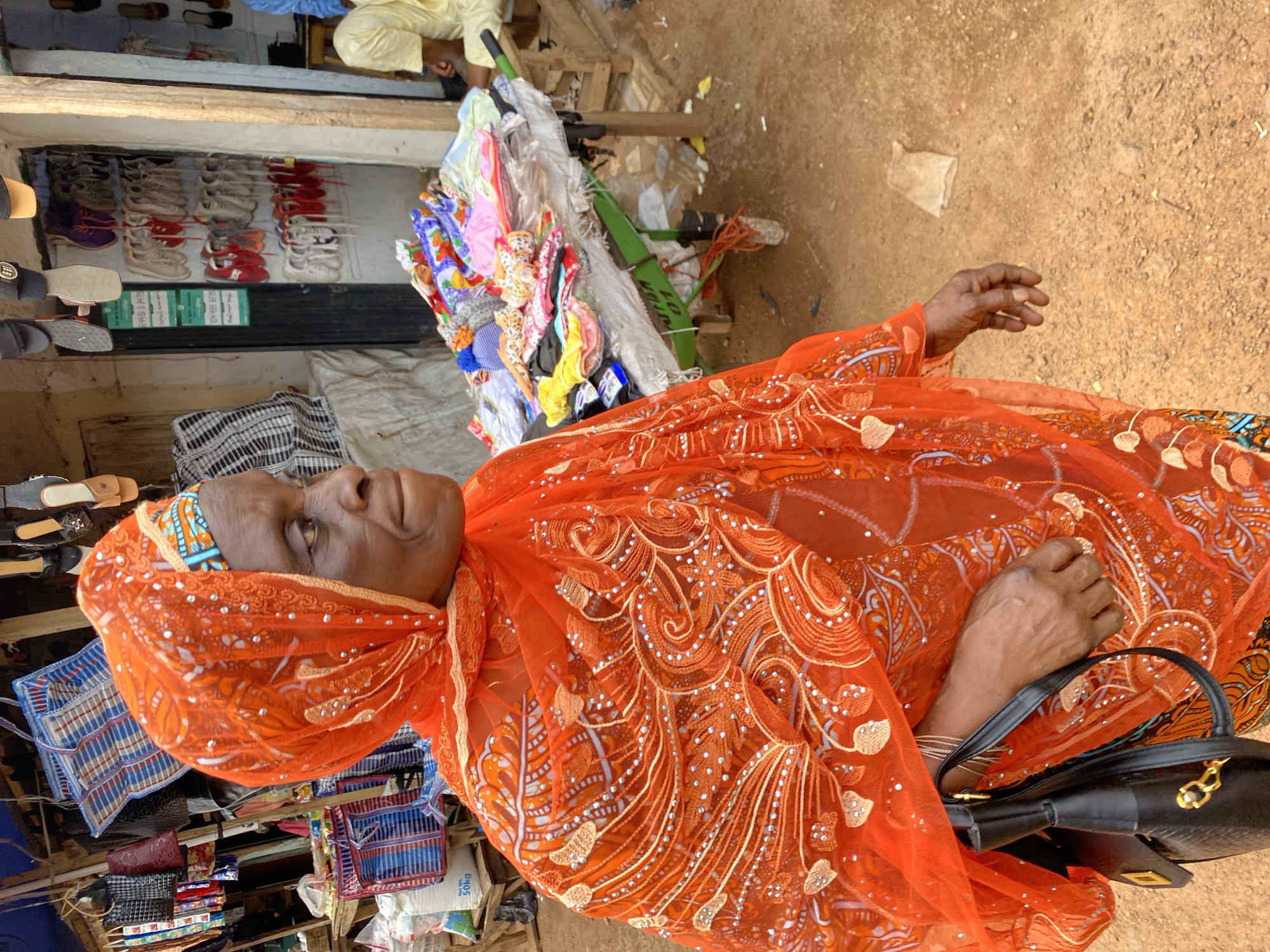 A woman, adorned in a vibrant orange headscarf and matching embroidered garment, is seen in an outdoor marketplace in Bukuru, Nigeria. She is positioned in the foreground, with her body turned to the right, looking away from the camera. Her left hand, holding a black handbag, is visible. Behind her, a stall displays various items, including colorful textiles and what appear to be shoes hanging from a frame. A person wearing a yellow garment is seated in the background to the right. The ground is dusty and appears to be unpaved.
