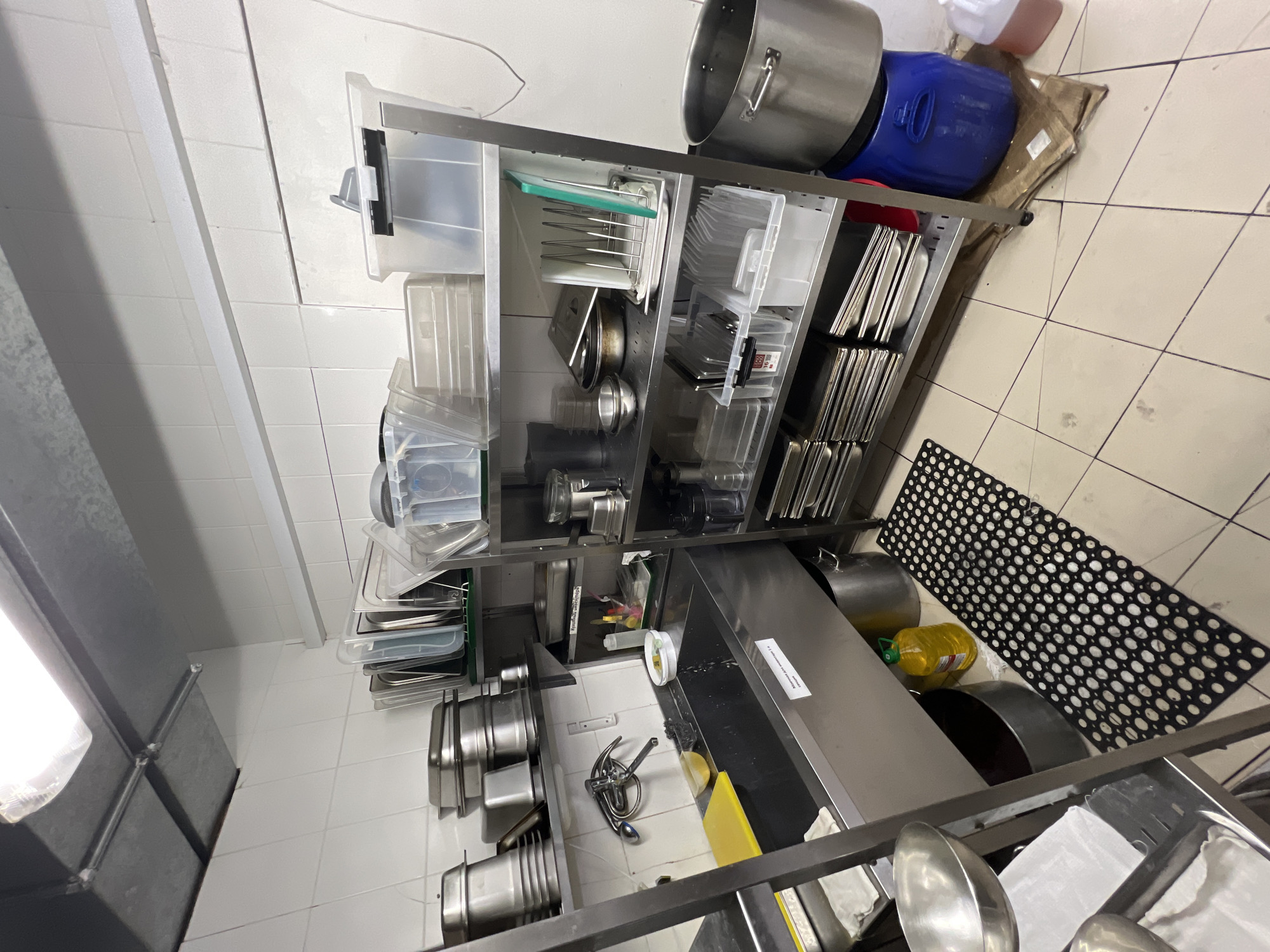 The image depicts a commercial kitchen environment, likely in an industrial or restaurant setting. The scene is dominated by stainless steel shelving units filled with various kitchen equipment, including stacks of metal pans, plastic containers, and bowls. A large stainless steel sink with a faucet is a prominent feature in the foreground, with a yellow cutting board and a sponge nearby. A black mat with circular cutouts is placed on the tiled floor in front of the sink. To the right, a large metal pot sits on a blue plastic container, and another large pot is visible on the floor. The walls are tiled in white, and the ceiling has overhead lighting. There are no people visible in the image, and no specific actions or interactions are depicted. The overall impression is one of a functional, working kitchen space. The location is identified as Plastunivska, Russia.