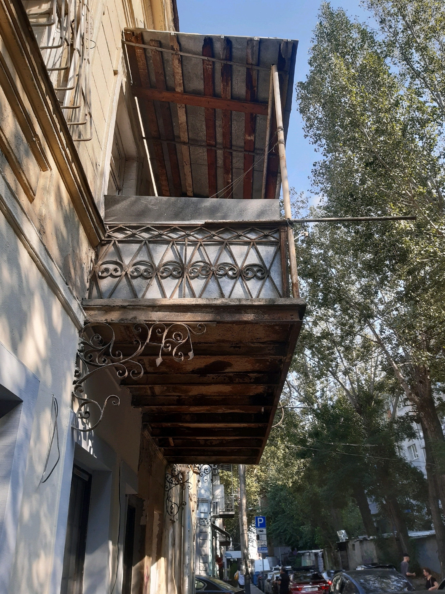 The image shows a weathered, wooden balcony with ornate wrought-iron railings extending from a building. The balcony's roof is made of corrugated metal sheets supported by wooden beams. Below the balcony, an entrance to the building is visible, with a dark doorway. The building's facade is a neutral color, showing signs of age and wear. A modern, dark-colored car is parked on the street in front of the building. Several trees with dense green foliage line the street, with their branches partially obscuring a clear blue sky. The scene appears to be a residential street in the city of Tbilisi, Georgia, characterized by its older architecture and ample greenery.