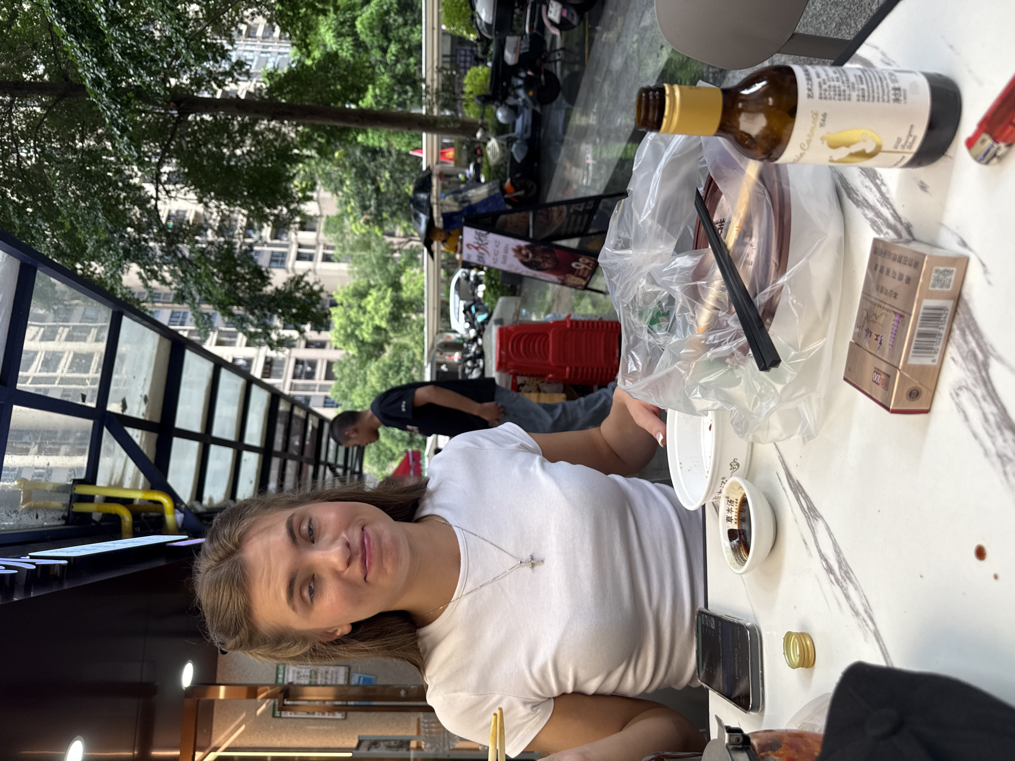 A young Caucasian female with light brown hair, wearing a white t-shirt and a silver cross necklace, is seated at a marble-topped table, holding chopsticks. She is making a grimacing expression. In front of her, on the table, are various items including a smartphone, a small dish of soy sauce, a bowl covered in plastic wrap containing food and chopsticks, a pack of cigarettes, a bottle of what appears to be wine, and a red lighter. The background shows a street scene with trees, buildings, and parked motorcycles. The scene appears to be an outdoor seating area of a restaurant in Haizhu District, China, with a covered walkway extending over the dining area.