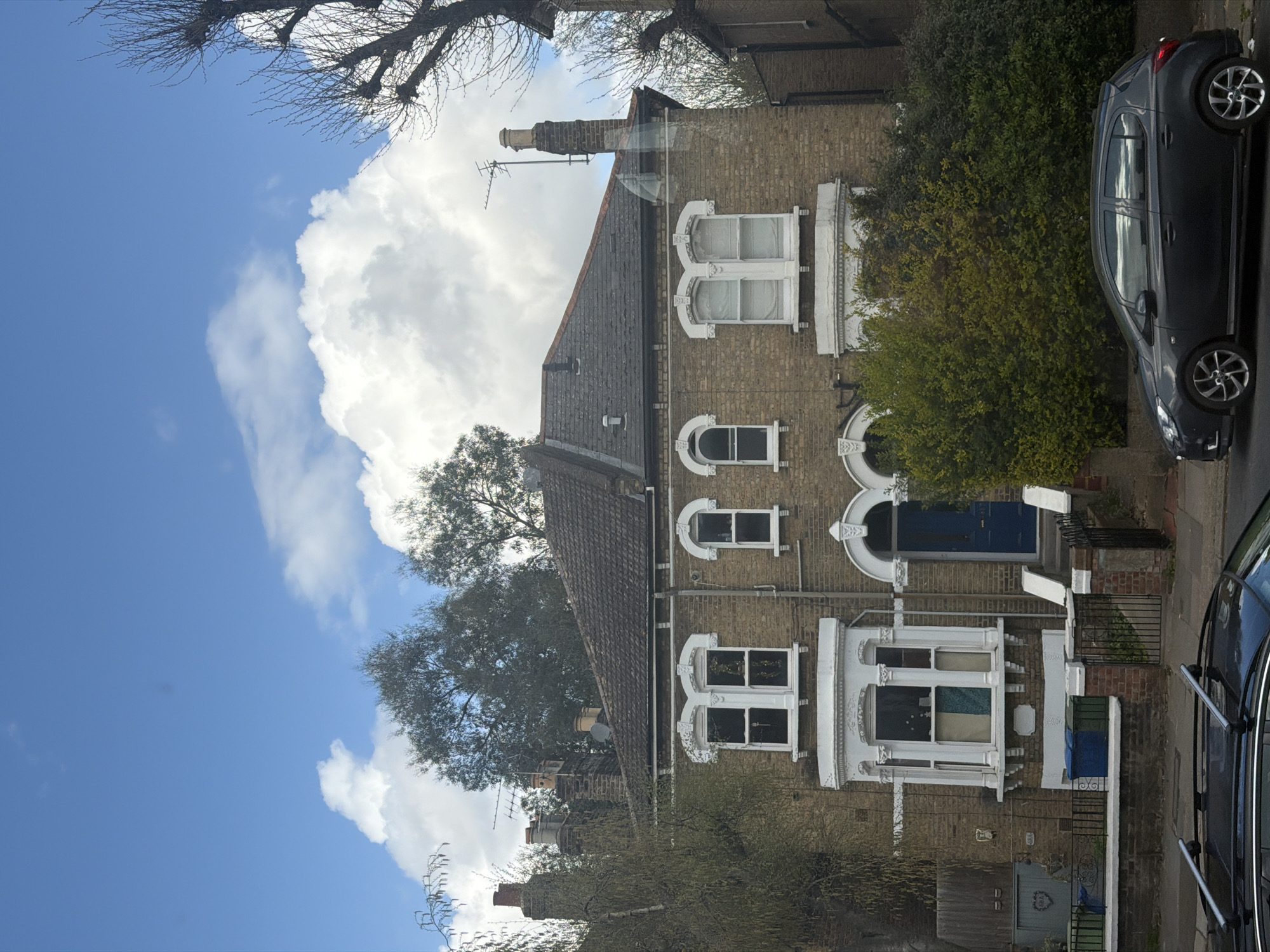 The image shows a residential street scene in London, United Kingdom, featuring terraced houses with brick facades. The houses have white-framed windows of varying styles, including arched and bay windows. Blue front doors are visible. Lush greenery, including trees and bushes, is present. A dark-colored car is parked on the street. A large, white cumulus cloud dominates the sky, contrasting with the blue sky. Branches from a bare tree are visible in the upper left corner. Chimneys and television aerials are on the rooftops.