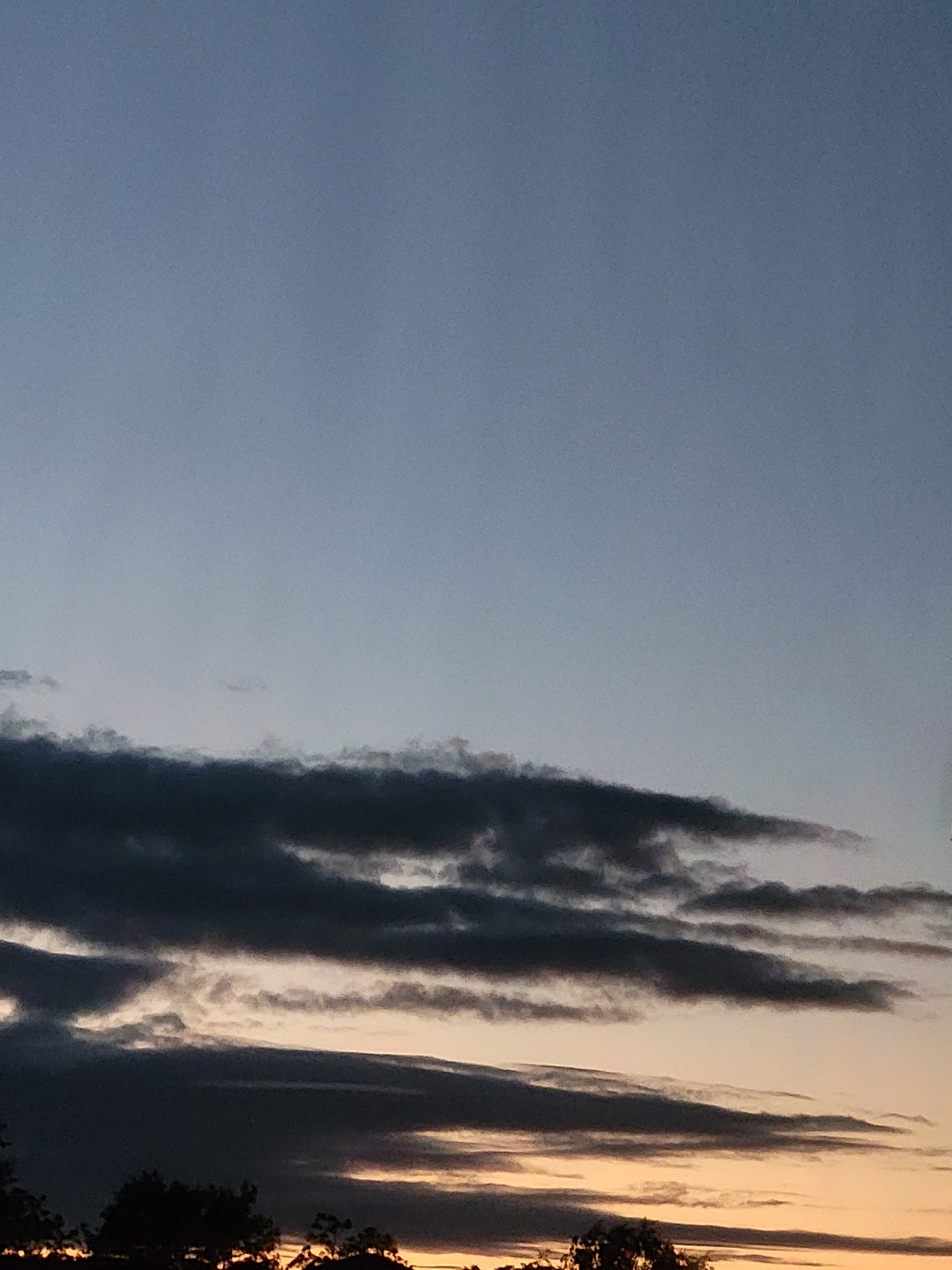 The image is a vertical, full-frame shot of a dusk sky, possibly taken in Ozolnieki, Latvia. The sky transitions from a pale, cool blue at the top to a warmer, orange hue near the horizon. Several layers of clouds are visible, with the lowest layer being the darkest and most distinct, appearing almost black and streaky, possibly indicating turbulent weather or the setting sun behind them. Above these dark clouds, lighter, wispy clouds are scattered, catching the fading light. The bottom edge of the frame shows the silhouetted tops of trees, their dark forms contrasting with the colorful sky. There are no people or specific objects identifiable in the image, focusing solely on the atmospheric conditions.