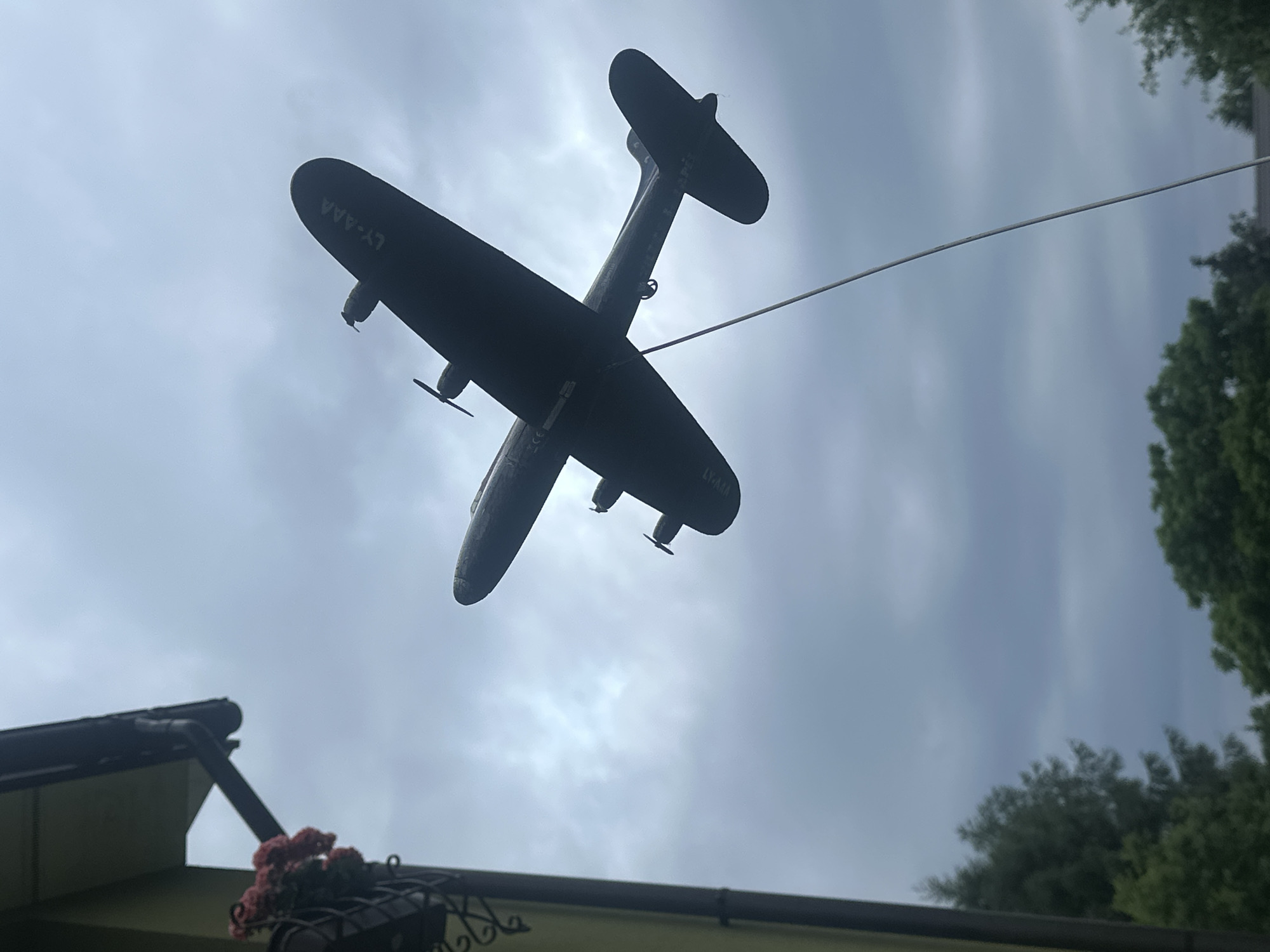 The image shows a silhouetted model airplane suspended in the air against a cloudy sky. The airplane is dark and appears to be made of plastic or a similar material. It has two propellers on its wings and a tail fin. A thin wire runs from the airplane to the right of the frame, suggesting it is being flown or displayed. The airplane has the registration "LY-AAA" visible on its fuselage. Below the airplane, a portion of a roofline and some greenery are visible, hinting at an outdoor setting. The presence of a roof and trees suggests this might be a residential area or a park. The sky is overcast, indicating a cloudy day, possibly with some diffused sunlight breaking through. No people are visible in the frame. The overall impression is of a whimsical display, possibly a toy or a decorative item. The location context indicates Lapės, Lithuania.