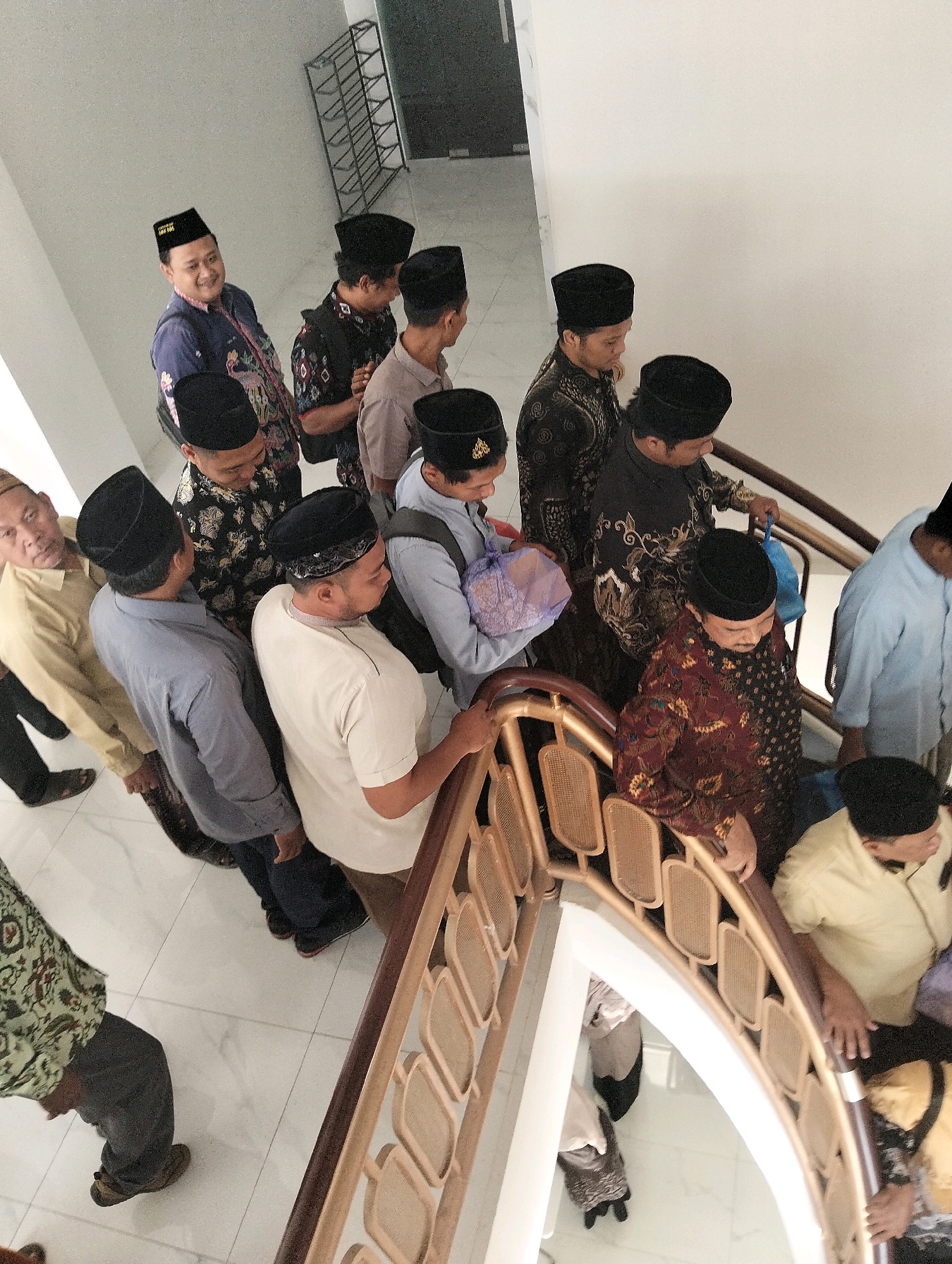 This is an overhead, high-angle, full shot of a group of at least ten adult males, most wearing traditional Islamic attire and skullcaps, ascending a curved, modern staircase inside a building. The individuals are positioned on the steps and landing, with some looking forward, others down, and one smiling toward the camera. The staircase features a light-colored, possibly marble floor and a light-colored wall. The banister is made of wood with rattan-like inserts. The lighting is bright and even, suggesting an indoor setting. The individuals are diverse in age and attire, with some wearing batik-patterned shirts and others plain shirts. Some are carrying small bags. The context suggests a gathering or event, possibly religious in nature, taking place in Paciran, Indonesia.