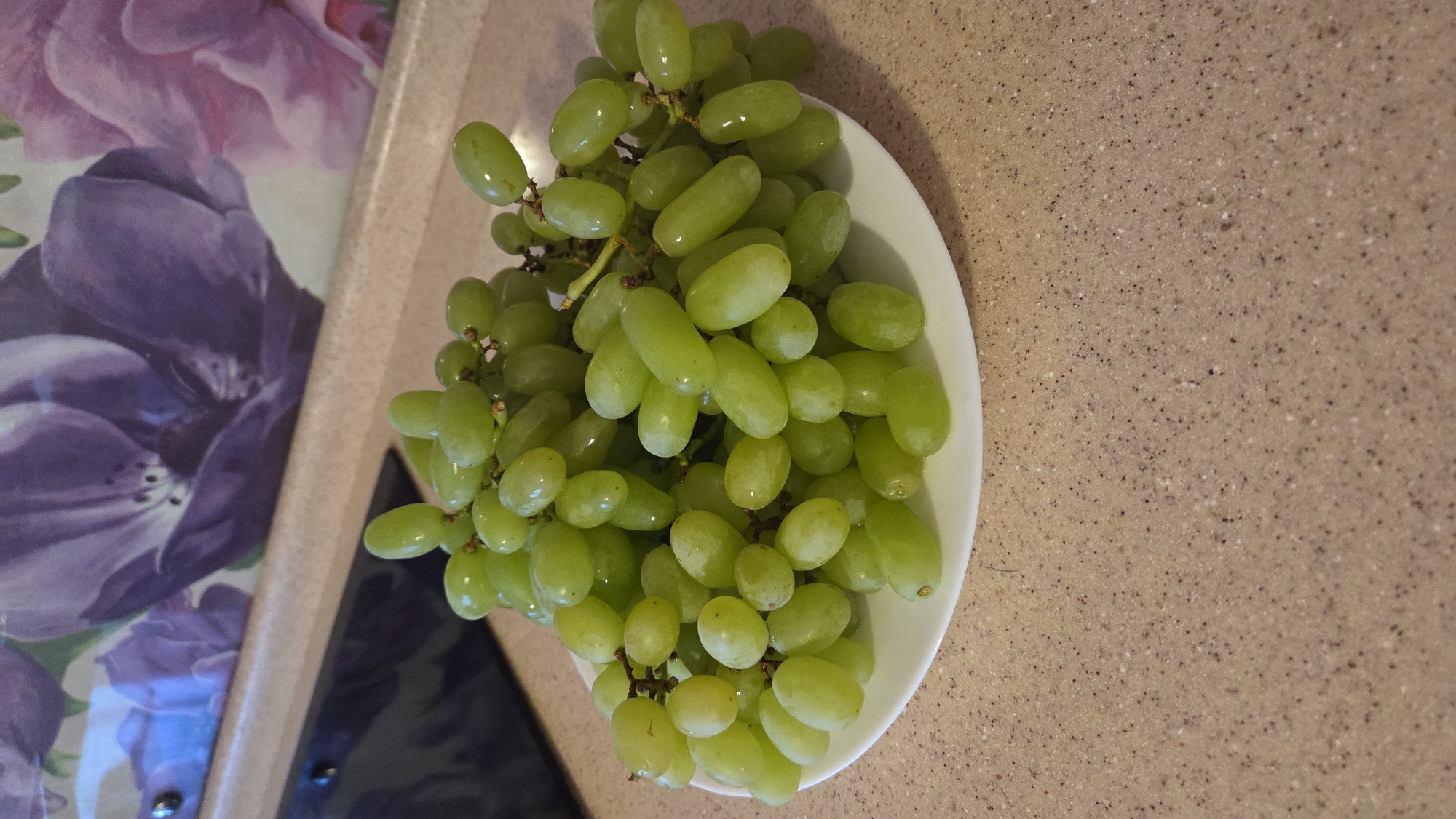 A generous cluster of fresh green grapes is prominently displayed on a white, oval-shaped plate or shallow bowl in a high-angle, close-up shot. The grapes are bright, light green, appearing plump and juicy, with several small water droplets visible on their skins, suggesting they have been recently washed. They remain attached to their stems, forming a natural bunch that largely fills the plate.
The plate of grapes rests on a light brown or beige countertop, which exhibits a subtle, speckled pattern. To the left of the grapes, a portion of a fabric item, likely a placemat or tablecloth, is visible. It features a vibrant floral design, dominated by various shades of purple and hints of pink, depicting large, soft-petalled blooms. A dark, reflective surface, possibly part of a kitchen appliance or a different object, can be seen beneath the floral fabric towards the bottom-left edge of the frame.
The setting appears to be an indoor domestic space, such as a kitchen or dining area. The lighting is even and bright, highlighting the freshness of the fruit. There are no people visible in the image, nor is there any discernible text or specific branding. The scene conveys a simple presentation of fresh fruit ready for consumption.