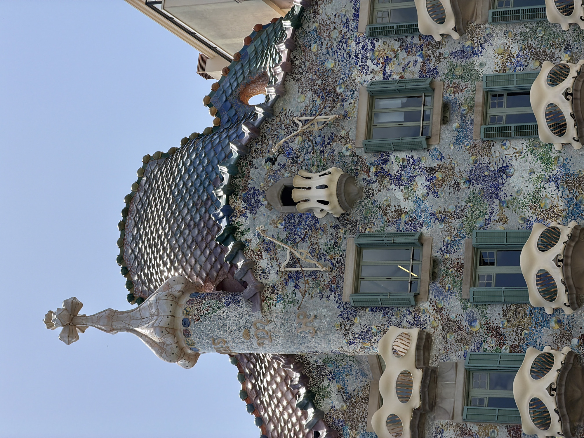 The image shows a close-up, low-angle view of Casa Batlló in Barcelona, Spain. The facade of the building is adorned with a mosaic of colorful tiles, predominantly in shades of blue, green, and white, creating a textured and organic appearance. Numerous decorative elements, including rounded balconies that resemble bone structures, stand out against the mosaic. Green shutters are visible on the windows, some of which are open. The roof features a unique, curved design covered in iridescent tiles that shimmer in the sunlight. A prominent turret topped with a decorative cross rises from the roofline. The sky is a clear, bright blue. The scene is devoid of people or any discernible activity.