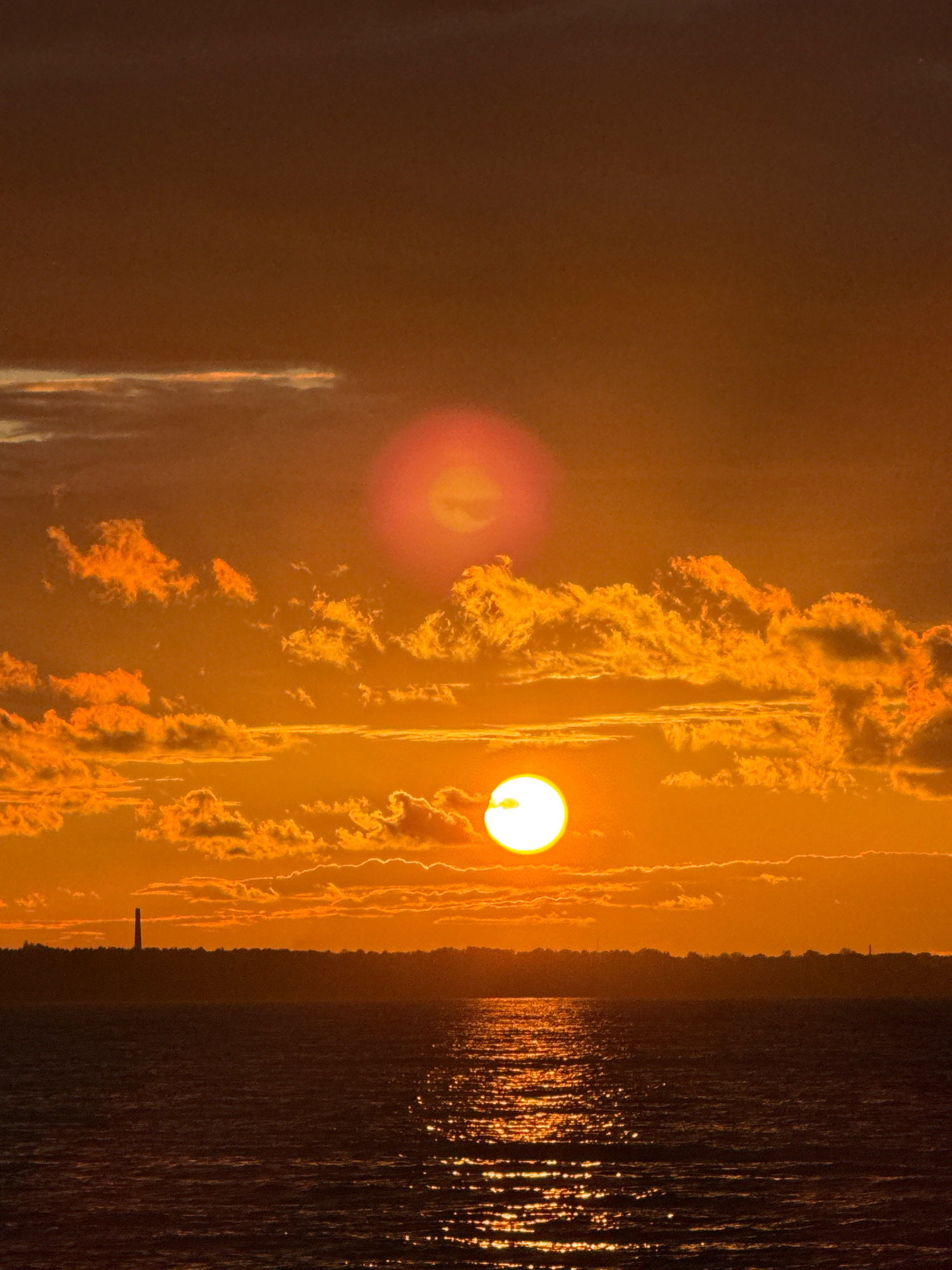 The image depicts a sunset over a body of water. The sky is a deep orange and brown, with several clouds illuminated by the setting sun. There are two visible suns, one larger and brighter in the lower half of the image, and a fainter, reddish disc higher up. The sun's reflection shimmers on the water's surface. A silhouetted landmass with a tall, slender structure resembling a lighthouse or tower is visible on the horizon. The scene is likely a coastal or lakeside area. There is no indication of the city of Limuzi, Russia in the image.