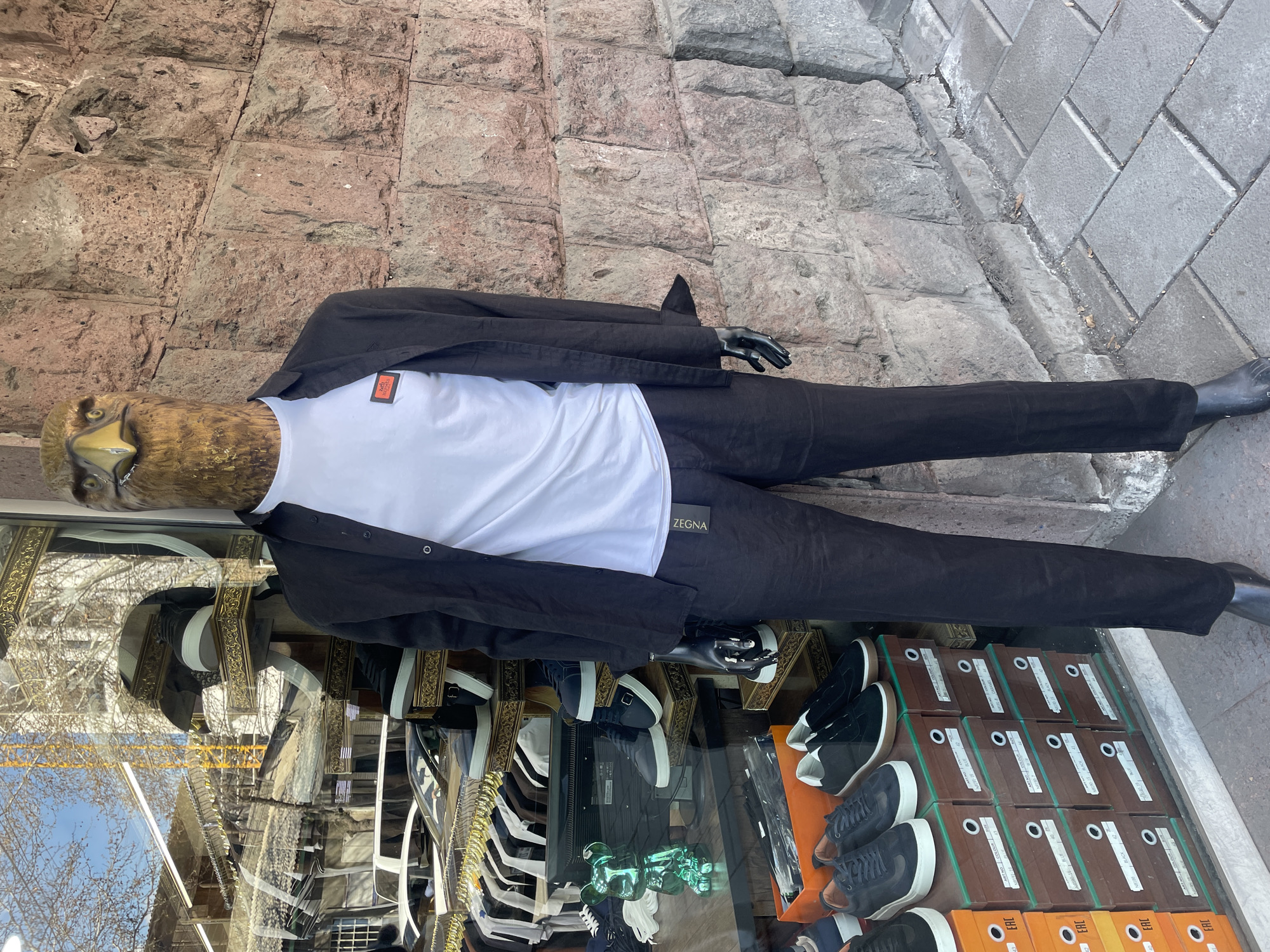 A mannequin with an eagle head stands in a shop window in Yerevan, Armenia. The mannequin wears a white t-shirt under a black jacket and black trousers, with black shoes. Reflected in the shop window, buildings and sky are visible. Inside the shop window, numerous shoe boxes and clothing items can be seen. The shop is situated on a paved street.