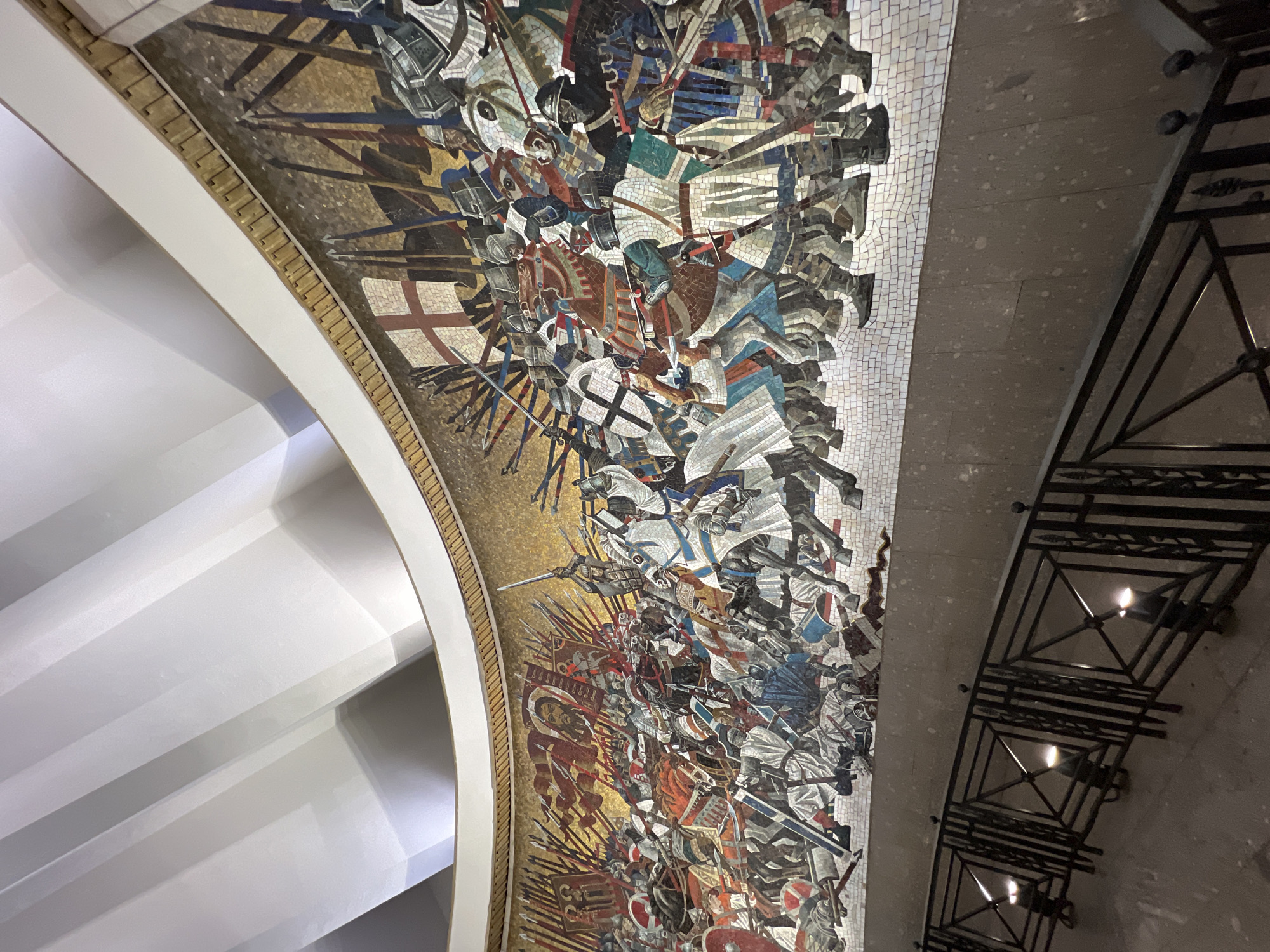 The image shows a mosaic on the ceiling of the Saint Nicholas Naval Cathedral in Saint Petersburg, Russia. The mosaic depicts a chaotic battle scene with numerous figures in armor and on horseback engaged in combat. Swords, spears, and flags are visible amidst the figures. The mosaic is composed of small, multi-colored tiles. Architectural elements, including white curved surfaces and a decorative trim along the edge of the mosaic, are also visible. A dark metal lighting fixture is present near the edge of the mosaic.