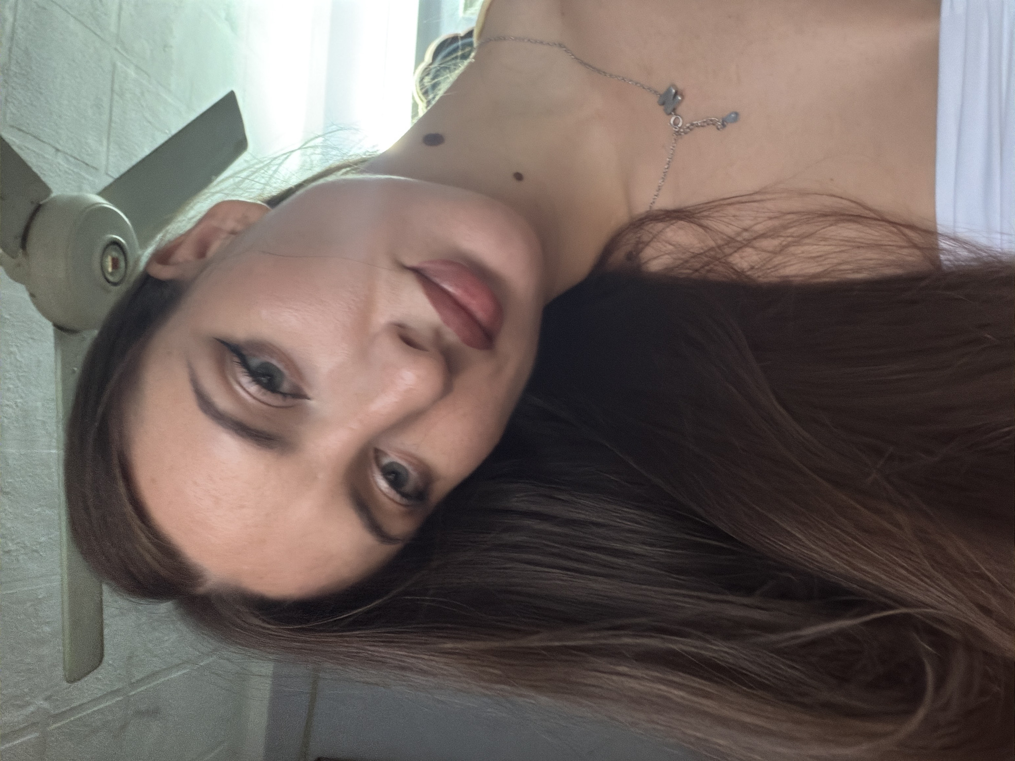 A close-up, overhead, indoor shot shows a young woman with long, brown hair. She is wearing a white tube top and a silver necklace with a small butterfly pendant. Her face is tilted to the left, and she has makeup on, including eyeliner and lipstick. Two dark moles are visible on her neck. The background includes a ceiling fan and a white ceiling. The image appears to be taken in a residential setting in 6th of October City, Egypt.