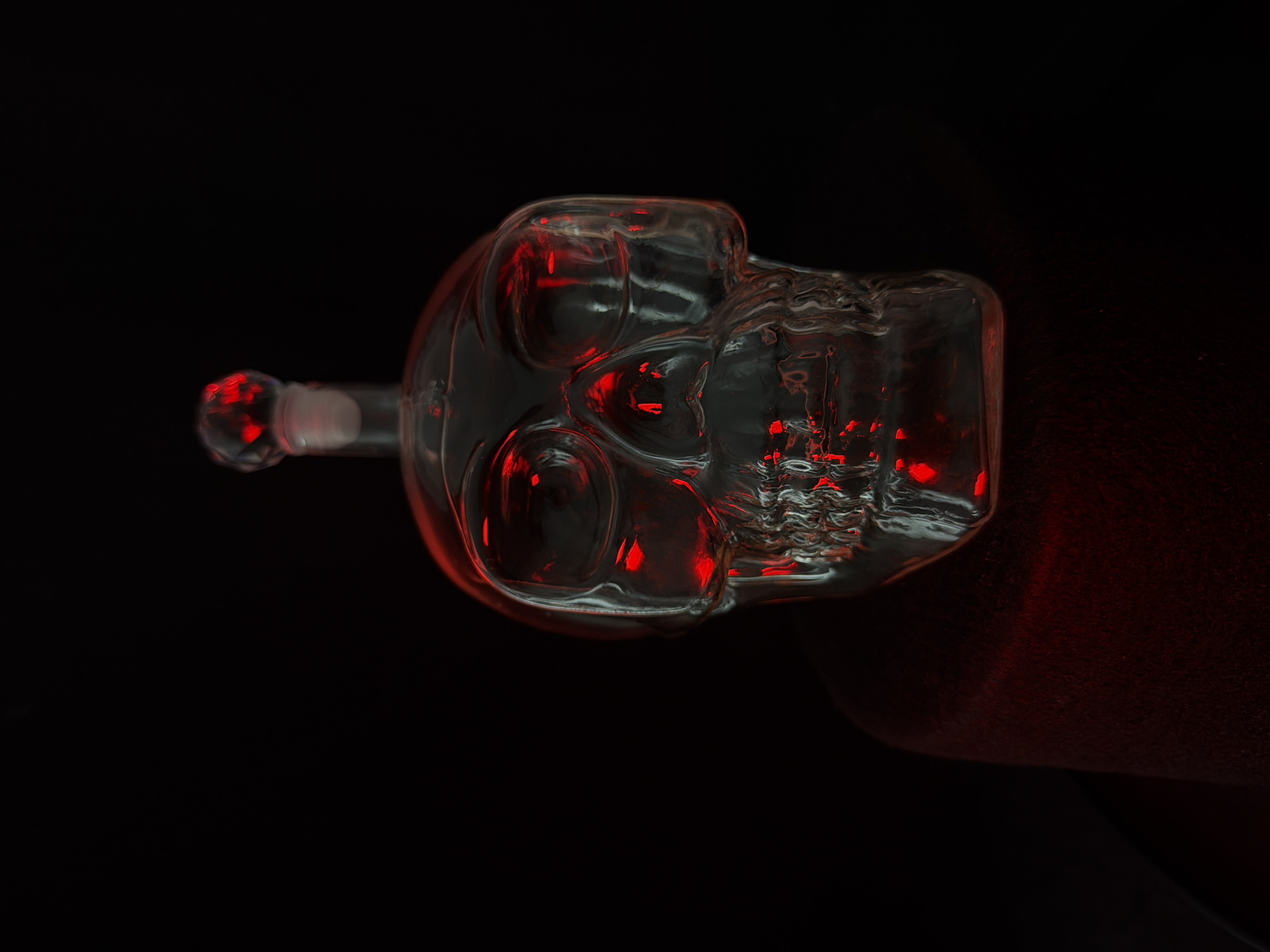 A glass skull-shaped bottle with red light glowing within it sits on a dark red, fuzzy surface against a black background. The bottle has a stopper at the top that is also clear glass with red light. The bottle is in focus and is the primary subject of the image. The red light reflects off the glass, creating an eerie effect. The overall mood of the image is dark and mysterious.