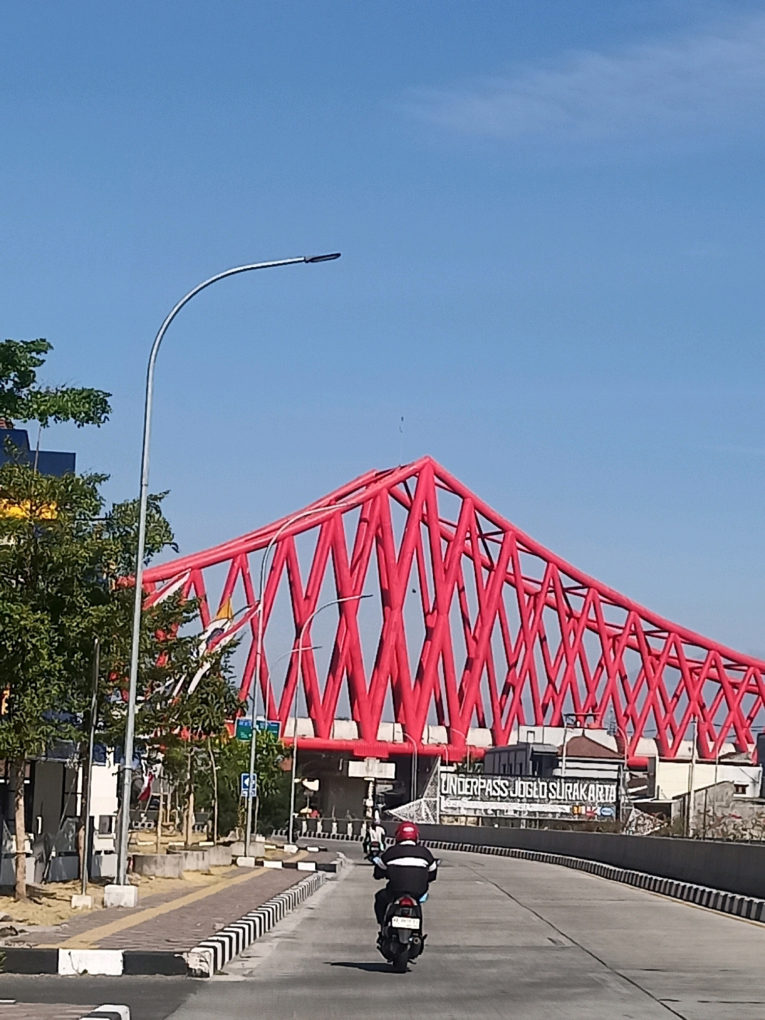The image shows a street scene in Surakarta, Indonesia, featuring a distinctive red truss bridge. A person riding a motorcycle with a red helmet is on the road, heading towards the bridge. Trees and streetlights are visible along the roadside. The bridge itself has a striking, geometric design, and a sign underneath it reads "UNDERPASS JOGLO SURAKARTA". The sky is clear and blue. The scene appears to be an urban environment on a sunny day.