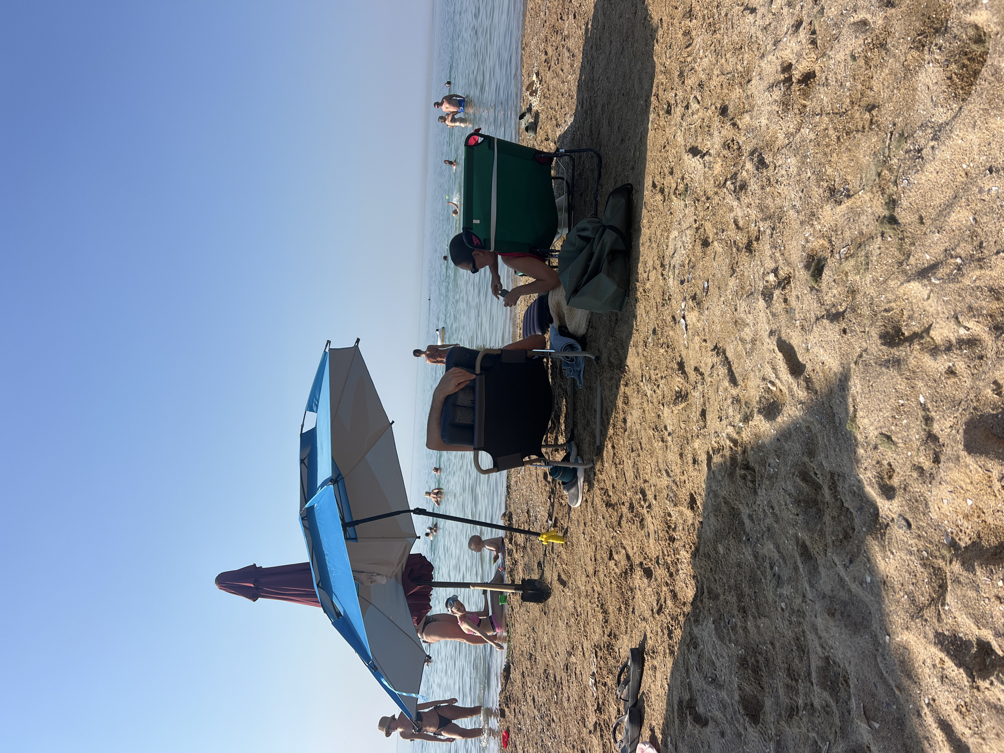 The image depicts a sunny day at a beach, likely Fontanka, Ukraine, given the context. The sky is a clear, bright blue, suggesting fair weather. People are engaged in typical beach activities. Some individuals are in the water, either swimming or wading, while others are relaxing on the sand.
In the foreground, two beach umbrellas are set up, providing shade. One is a large blue and white umbrella, partially obscuring a maroon umbrella. Beneath the blue and white umbrella, a person is sitting in a chair, and another person is partially visible. Further back, a green beach chair is occupied by a person wearing sunglasses and a red top, who appears to be looking at their phone. Next to this chair, a green bag rests on the sand. Other people are scattered throughout the scene, with several visible in the shallow water. The sand appears dry and textured, with shadows indicating the position of the sun. The overall mood seems relaxed and leisurely, consistent with a day at the beach. No visible text is present in the image.