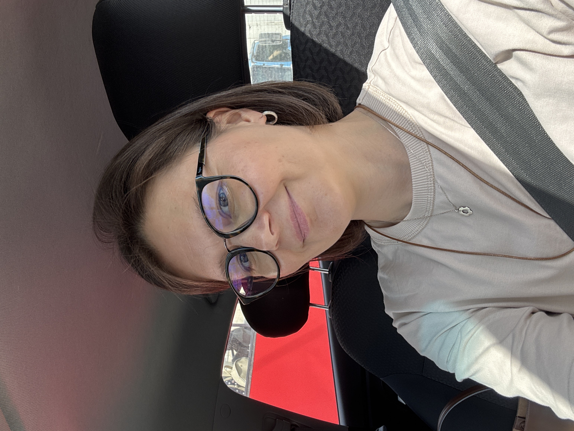 A woman wearing glasses and a seatbelt sits in a car. Outside the car window, a red surface is visible. The location appears to be Izhevsk, Russia.