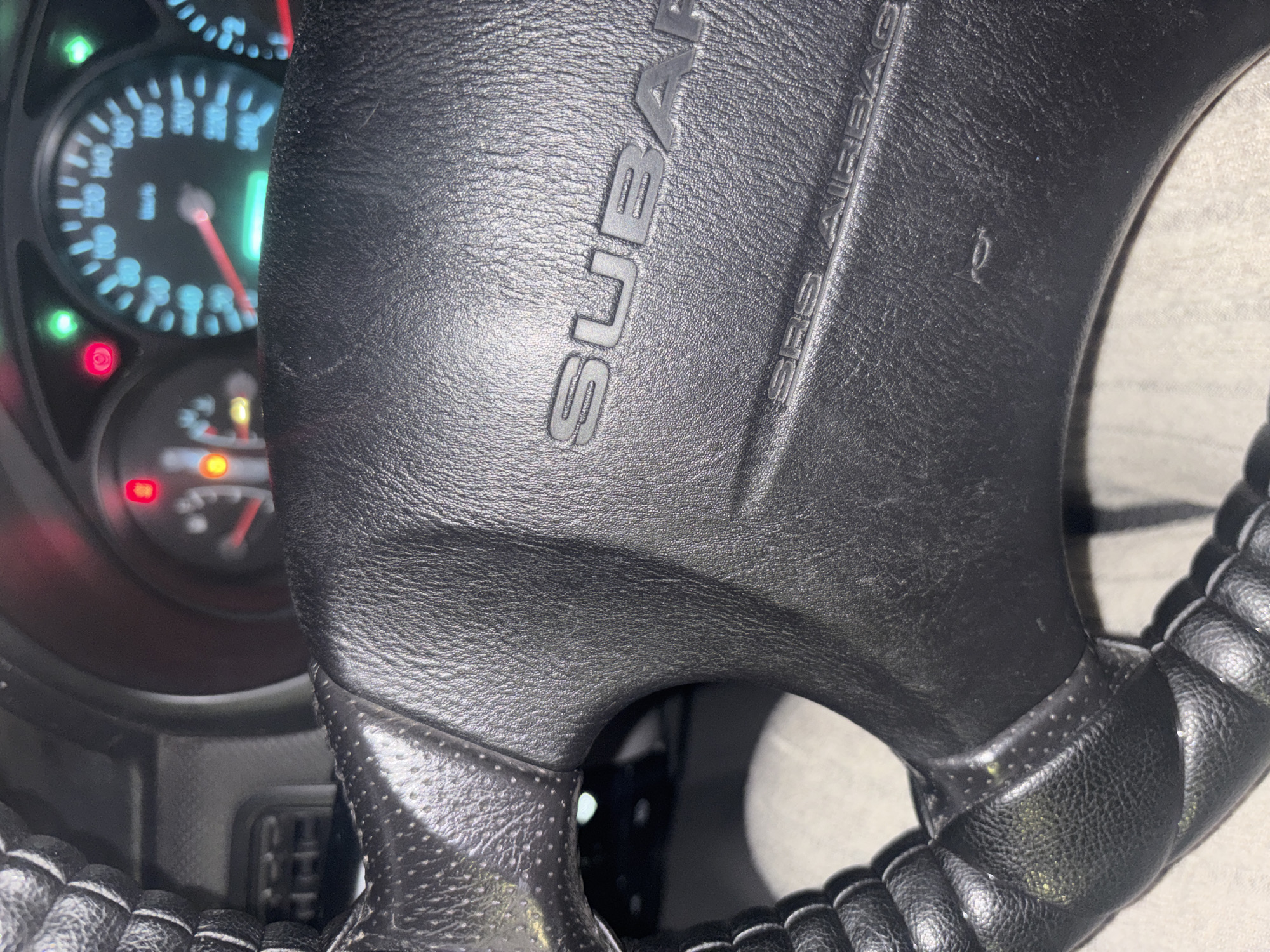 The image displays the interior of a vehicle, specifically focusing on the black leather steering wheel and a portion of the illuminated instrument cluster, located in the city of Dimitrovgrad, Bulgaria. The steering wheel, fitted with a ribbed, possibly aftermarket, grip on the left side, features a central airbag cover embossed with the word "SUBARU" at the top and "SRS AIRBAG" below. A small scuff mark is visible on the lower right quadrant of the airbag cover. Yellowish particulate matter is scattered across the steering wheel's textured surface, particularly on the left grip. Behind the steering wheel, the instrument cluster is partially visible, showing a speedometer marked in kilometers per hour (km/h) with a maximum reading of 240 km/h and the needle resting at 0 km/h. Multiple indicator lights are illuminated: a red circle with a 'P' symbol, a yellow circular warning light, a red warning light with an exclamation mark within a circle, and two green lights positioned at the top left and right of the speedometer, along with another green light at the bottom center of the cluster. The surrounding dashboard material is dark gray or black.