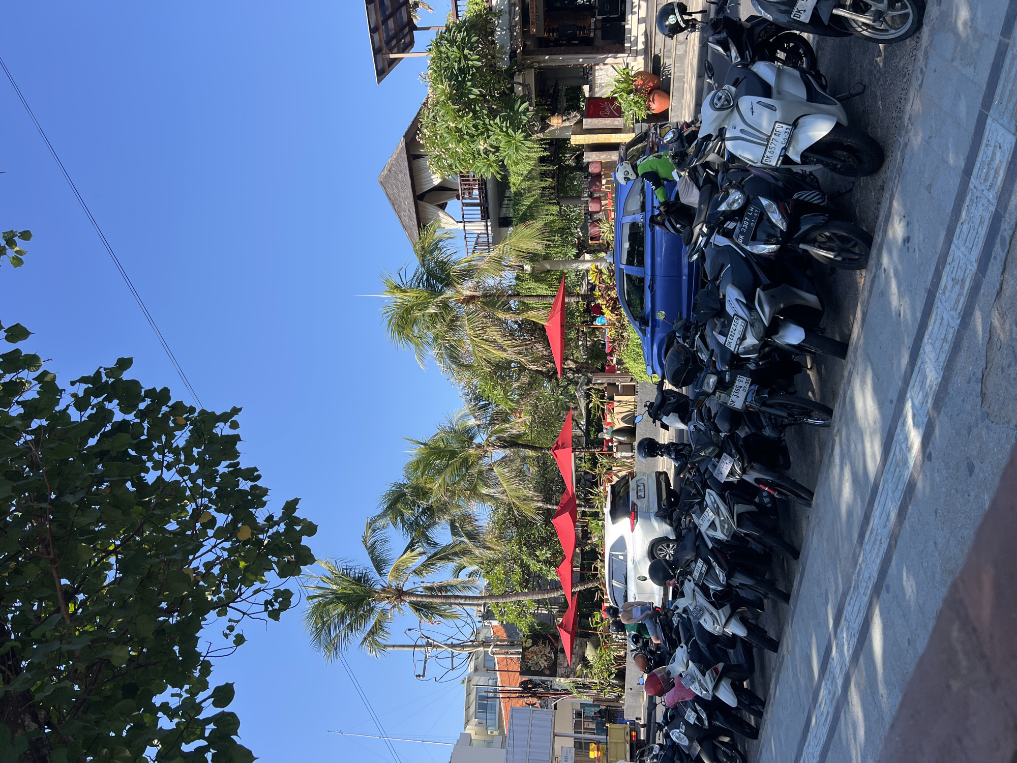 The image depicts a street scene in Kuta, Indonesia, on a clear, sunny day. In the foreground, a row of parked motorcycles and scooters lines a sidewalk. Behind them, a white SUV and a blue car are parked on the street. Beyond the vehicles, a building with an open-air upper floor featuring a pitched roof and draped curtains is visible, surrounded by lush greenery and palm trees. Several red umbrellas are set up in front of an establishment, suggesting an outdoor seating area for a restaurant or cafe. The sky is a vibrant blue, with some power lines and a few leaves from an overhanging tree visible at the top of the frame. A few individuals can be seen on the motorcycles and near the building.