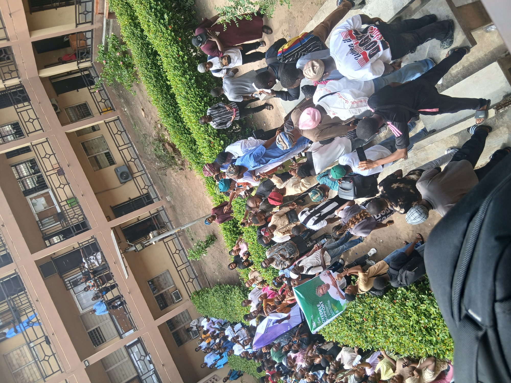 A large group of people, estimated to be over 100, is gathered outside a multi-story building in what appears to be a campus setting in Lagos, Nigeria. The building is light brown with dark brown window frames and railings. Some individuals are visible on the balconies of the building. The people are predominantly of African descent, and many appear to be young adults. Some are standing, while others are sitting or lying on the ground. There is a green hedge separating the crowd from the building. Several banners are visible within the crowd, including one with a portrait on it. Some people are looking down, and others are looking toward the direction of the banner. The ground is a mix of dirt, concrete, and greenery. A black backpack is visible in the lower right corner of the frame.
