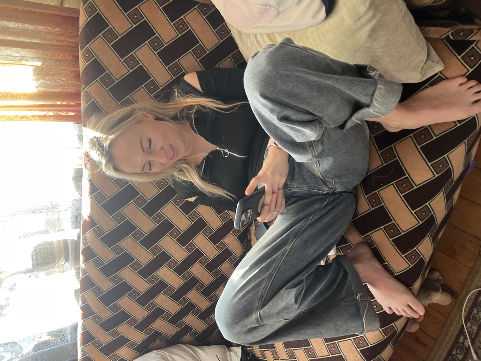 A woman with blonde hair is lying on a patterned brown and tan couch, looking at a smartphone. She is wearing a black zip-up shirt and blue jeans. A white pillow is behind her head. Her bare feet are visible on the right side of the image. A window is visible on the left side of the frame. The location in Podolsk, Russia is not indicated.