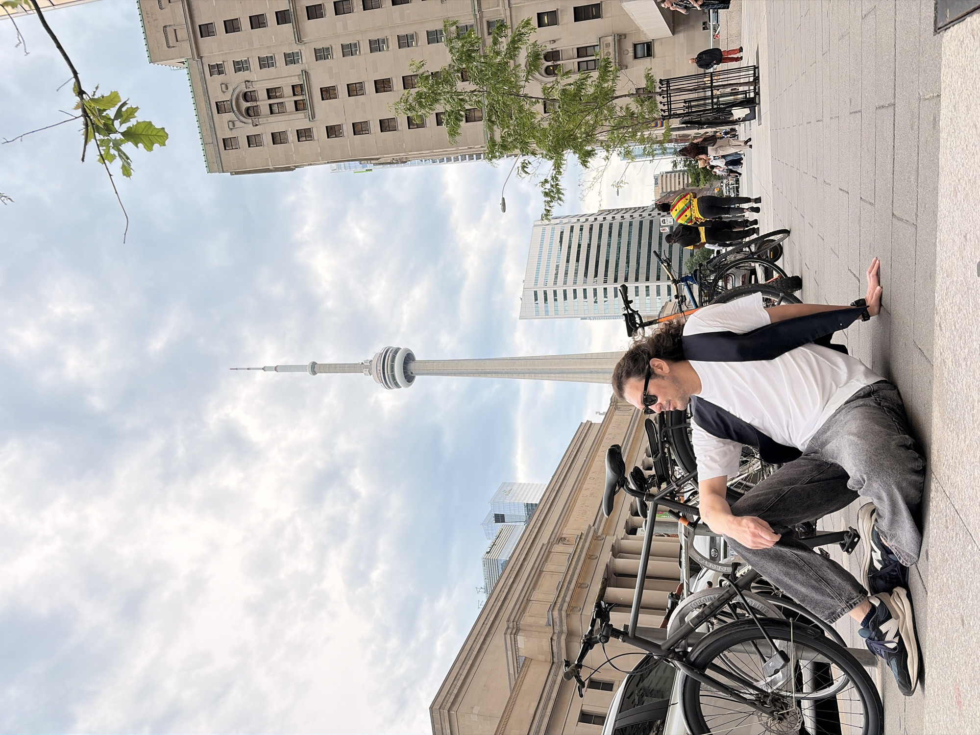 A man with long, dark, curly hair and sunglasses is sitting on the sidewalk in Toronto, Canada. He is wearing a white t-shirt, a dark vest, and distressed grey jeans. He has on dark sneakers with white soles. Two bicycles are parked next to him.
In the background, the CN Tower, a prominent landmark in Toronto, is visible. Several buildings are also present, including what appears to be Union Station with its classical columns, and modern glass skyscrapers. A few other pedestrians are walking on the sidewalk in the distance. The sky is cloudy. The man is looking to his right.