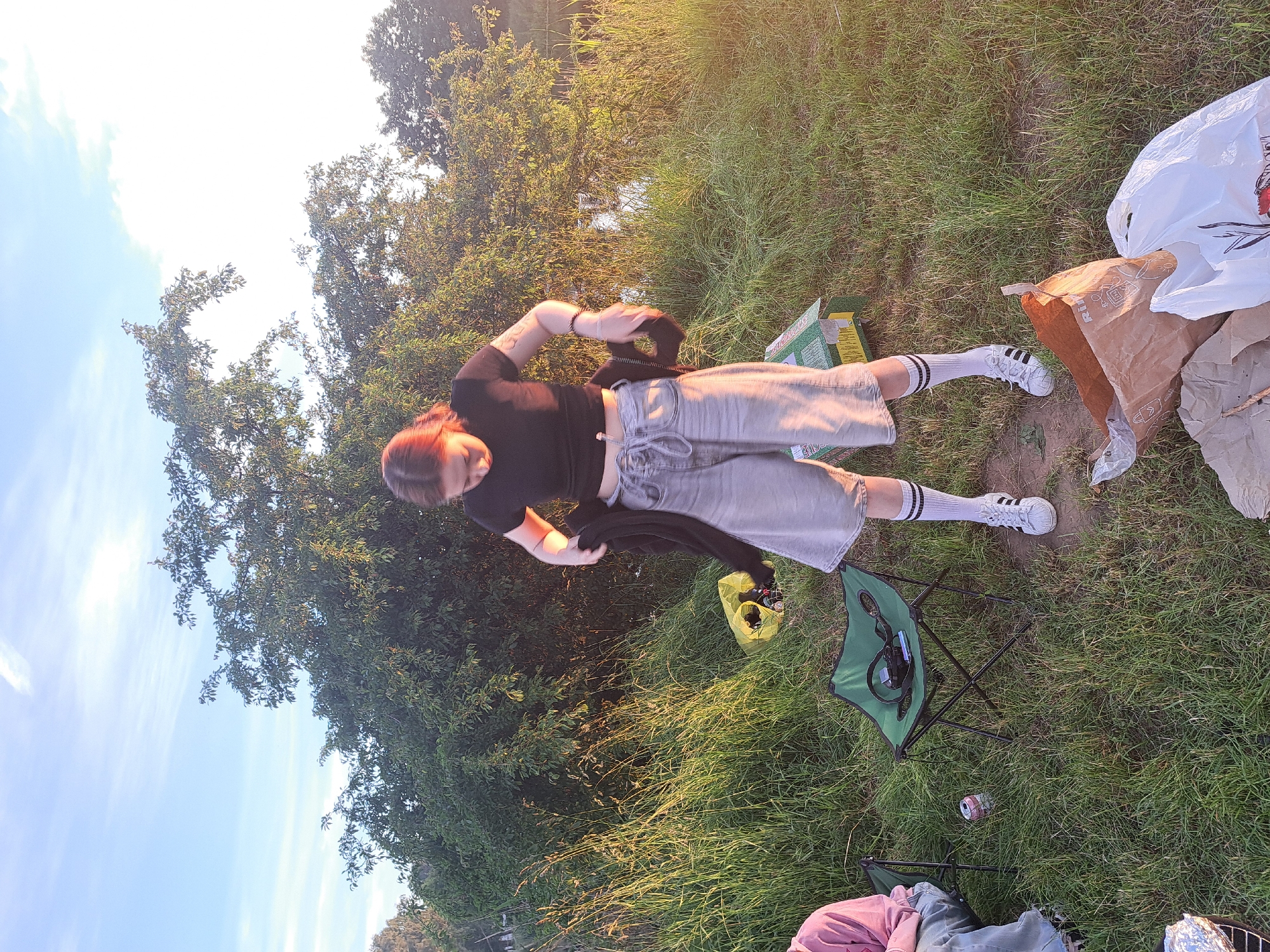 The image depicts a person, likely female based on visible features and clothing, standing in a grassy area with trees in the background. The person is wearing a black t-shirt, denim shorts, and white athletic socks with black stripes, paired with white sneakers. They are holding a black piece of clothing, possibly a jacket or sweater, in their hands and appear to be in the process of adjusting it or putting it on.
The scene suggests an outdoor setting, possibly a park or a campsite, given the presence of a green folding chair and other assorted items like bags and a beverage can on the ground. The lighting indicates it is either early morning or late afternoon, with the sun casting a warm, golden glow on the grass and trees, suggesting favorable weather. The sky is a clear blue with some wispy clouds visible.
The overall mood seems relaxed and casual, consistent with an outdoor leisure activity. While no explicit text is clearly legible on any of the objects, some items might contain branding or labels that are not discernible in the provided image. The presence of a can and camping gear suggests a recreational outing. The location context provided (Riga, Latvia) helps to place the scene in a specific geographical region.