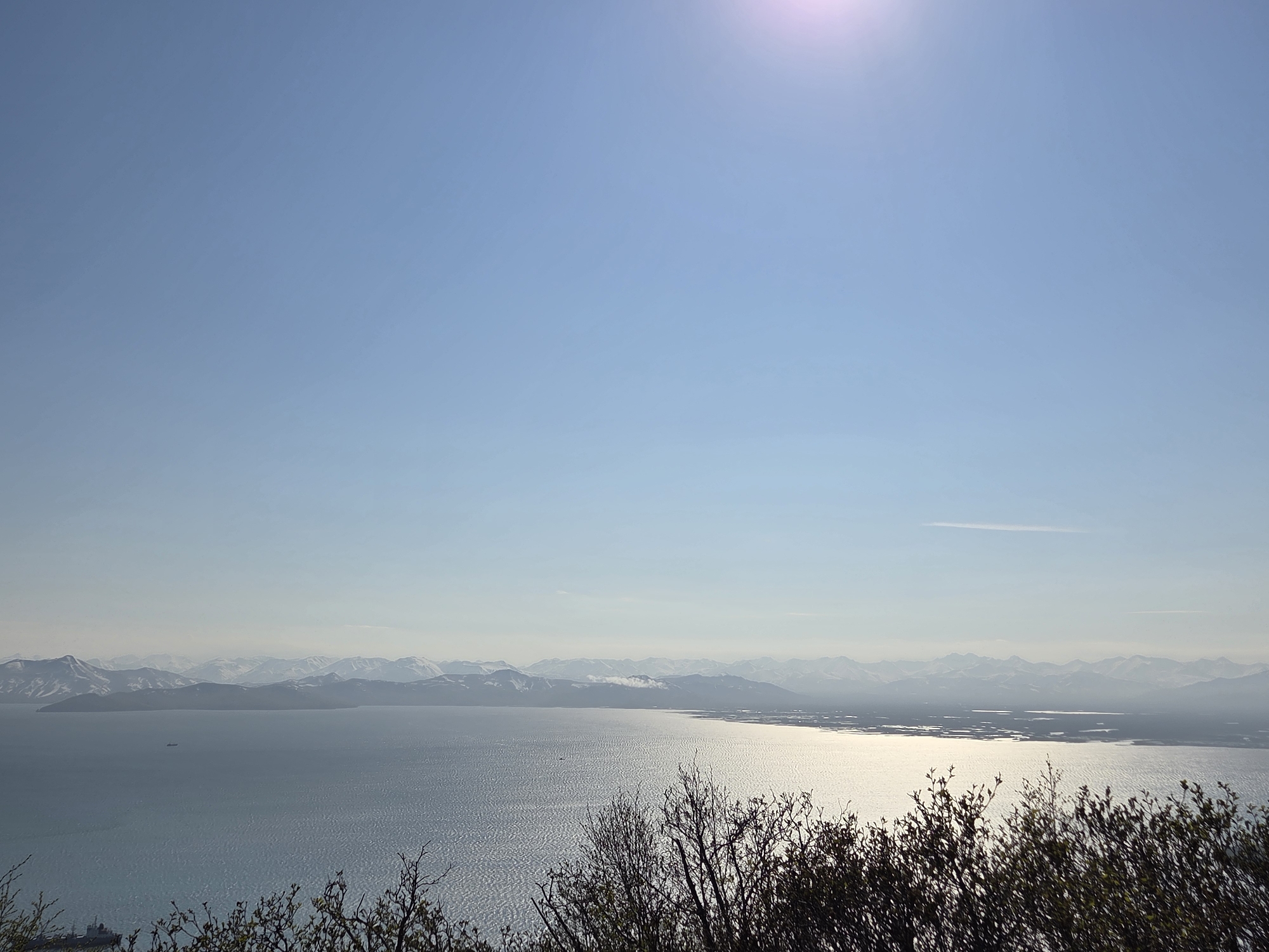 The image presents a wide-angle panoramic view of a serene body of water, likely a bay or sea, under a clear, bright sky with the sun visible as a lens flare in the upper right quadrant. In the distance, a range of mountains, capped with snow, forms a majestic backdrop. The water's surface is rippled, reflecting the sunlight in a shimmering pattern. In the foreground, the silhouettes of bare-branched trees and shrubs are visible, framing the expansive landscape. A small ship or boat is discernible on the water, and another larger vessel appears near the lower left edge of the frame. The scene suggests a natural, possibly coastal environment.