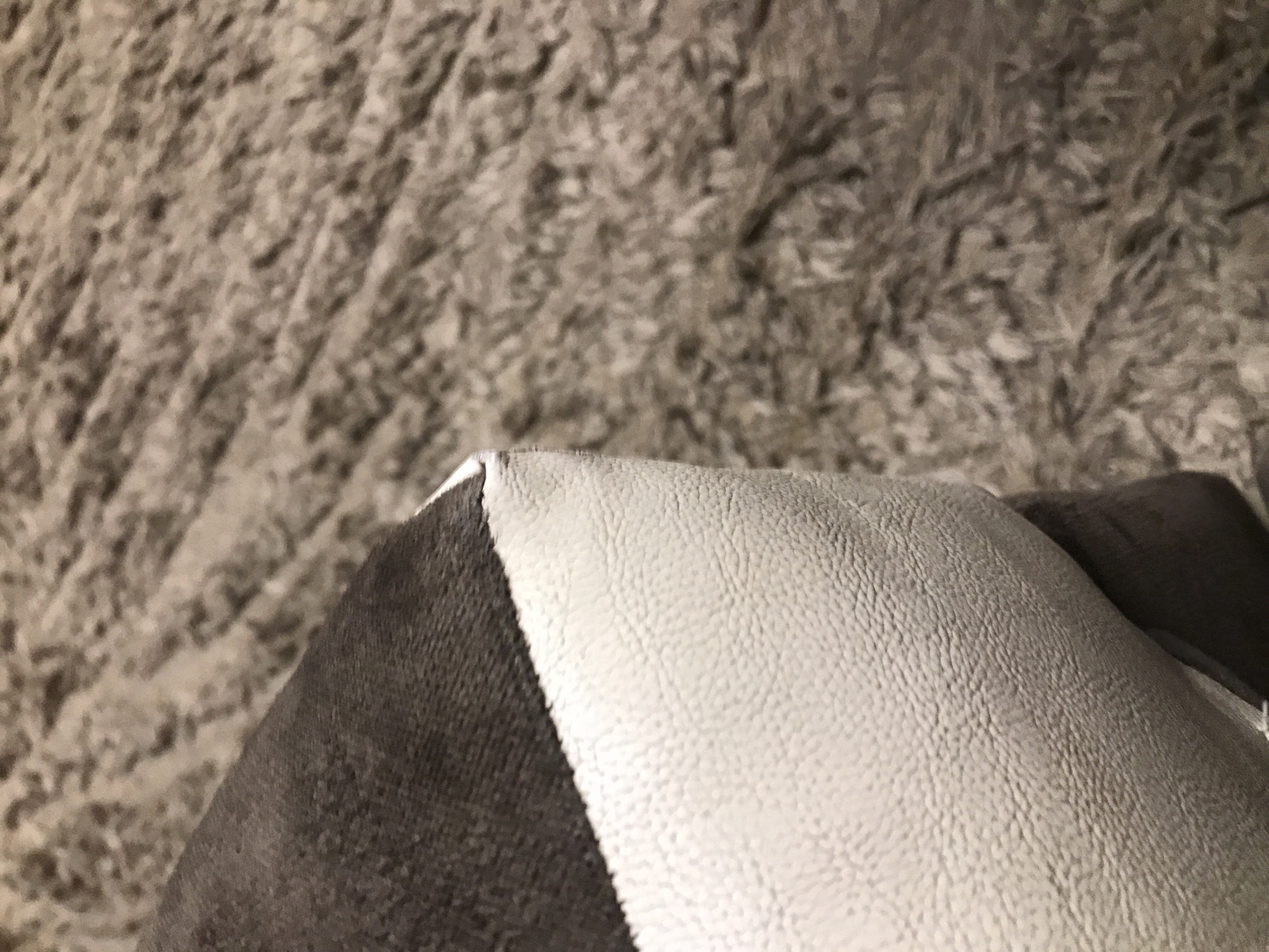 A close-up shot shows a section of a two-toned cushion resting on a textured gray surface, likely a rug. The cushion is divided into a dark gray velvety side and a light gray textured side, possibly imitation leather. The composition is tightly framed, focusing on the textures and materials. The location is unknown and cannot be determined from the visual data.