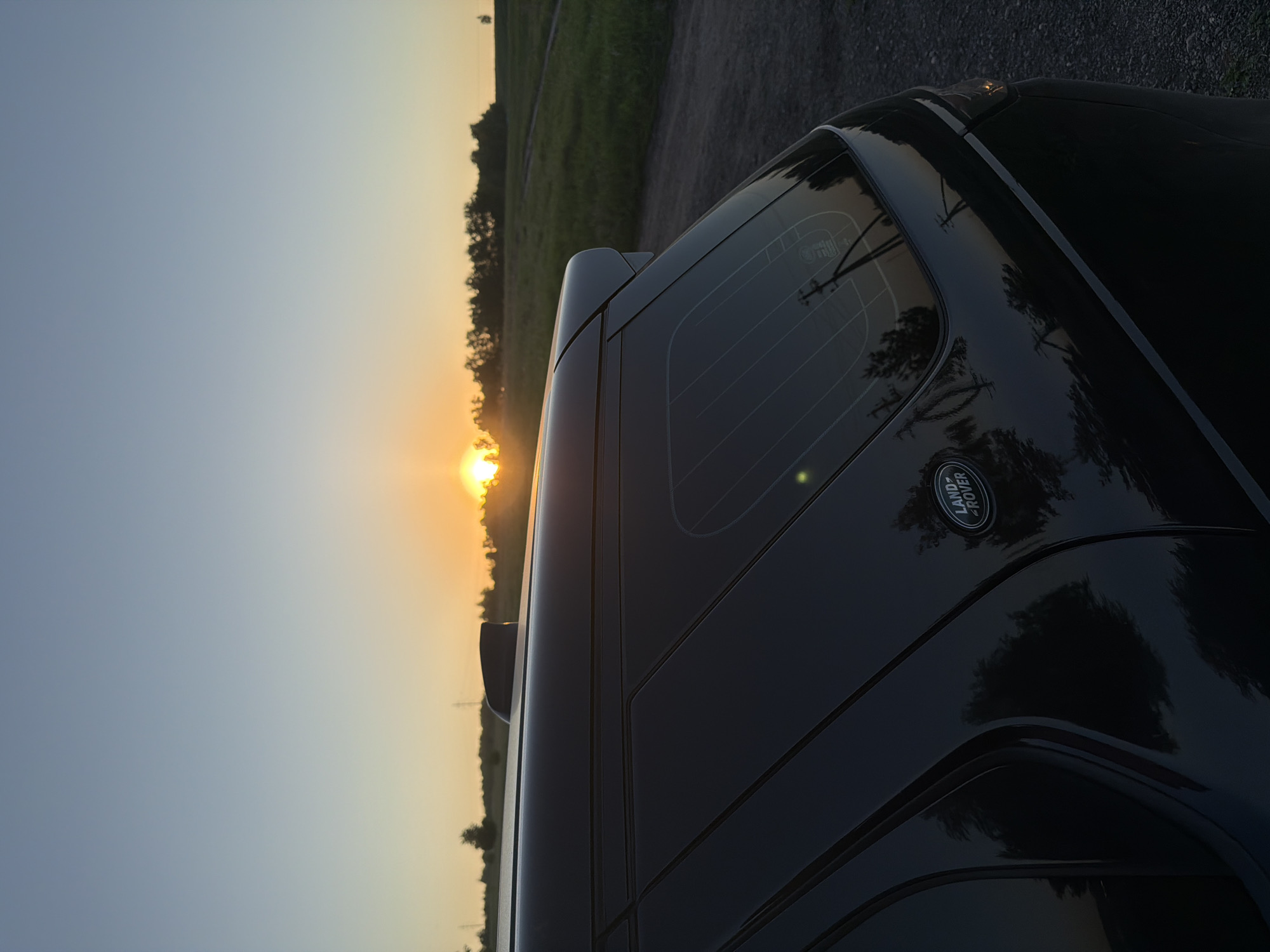 A low-angle, vertically oriented photograph captures the rear passenger side of a dark Land Rover vehicle against a vivid sunset. The sun, a brilliant golden sphere, is positioned low on the horizon to the left, bathing the scene in a warm, intense light. The sky above transitions from bright orange and yellow near the horizon to a softer blue-grey overhead.
A line of trees and bushes forms a dark silhouette against the glowing sky and sun, beyond which lies what appears to be a field or open landscape. The vehicle is parked on a dirt road or unpaved path. The glossy black surface of the Land Rover reflects the surrounding environment, with visible distortions of trees and the sky. The rear window displays characteristic defroster lines.
A clear "LAND ROVER" emblem is visible on the lower right portion of the vehicle's rear. The image evokes a serene and peaceful atmosphere typical of the golden hour, set in a natural or semi-rural environment, consistent with areas found near Krasnoyarsk, Russia. No individuals are present in the image.