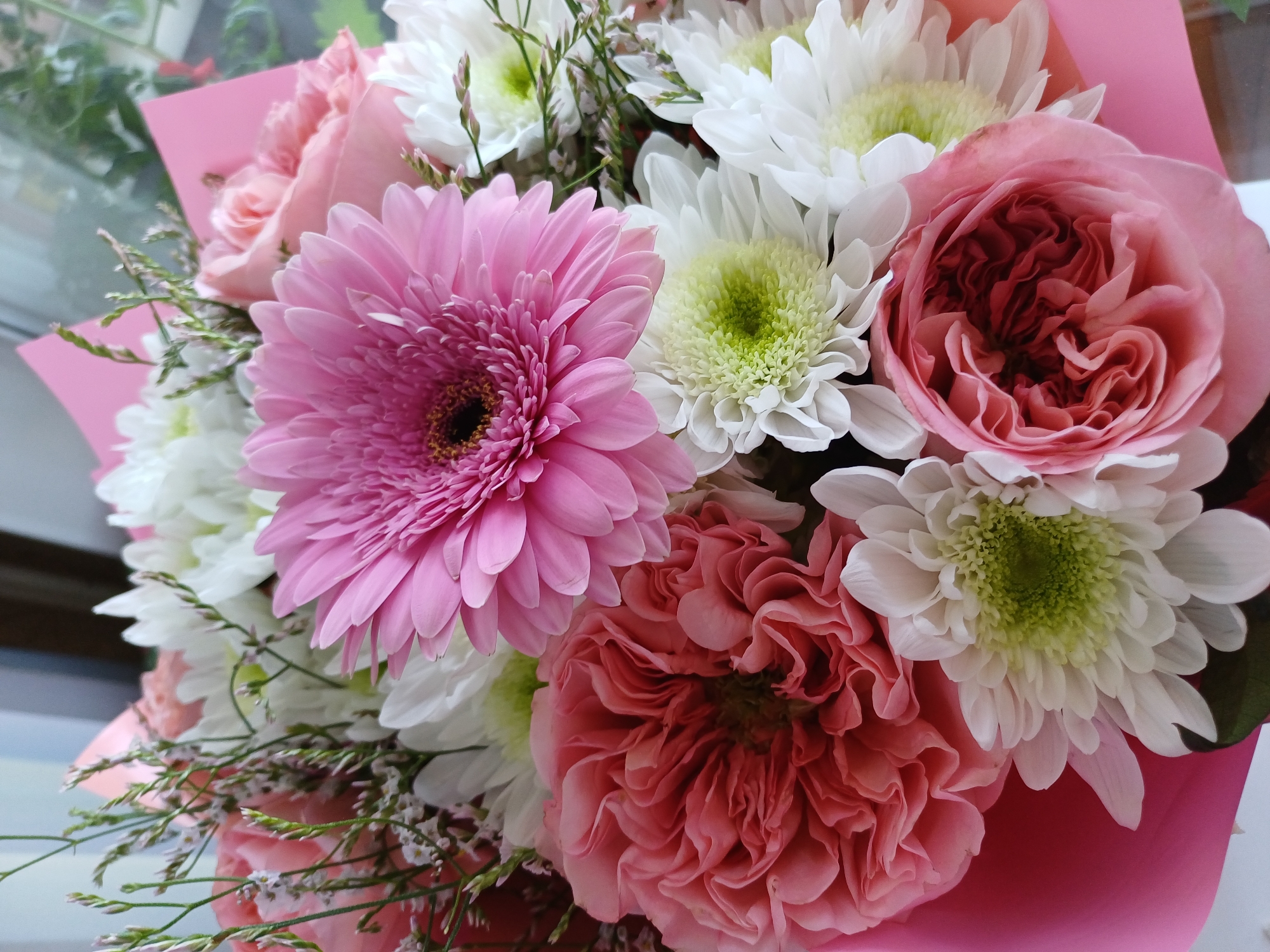 A close-up, overhead shot features a bouquet of flowers. Dominating the center of the frame is a large, vibrant pink Gerbera daisy with a dark center. Surrounding it are several white chrysanthemums with light green centers. Two large, ruffled pink roses are also prominent, one on the left side of the bouquet with deep pink petals and another on the lower right with lighter pink, tightly wound petals. Delicate sprigs of green filler flowers and thin, wispy green branches are interspersed throughout the arrangement. The bouquet is partially wrapped in a bright pink paper. The background is blurred, showing hints of a window and green foliage, suggesting an indoor setting near a window. There are no people or animals present. The location is unknown.