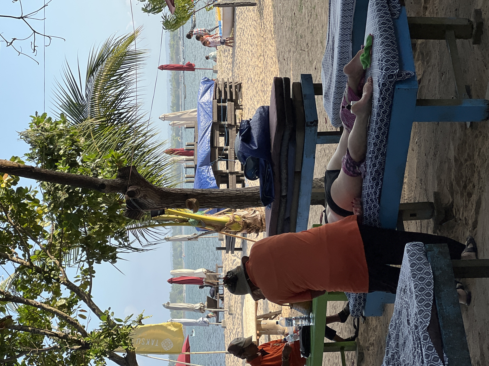 In Denpasar, Indonesia, on a sandy beach, a masseuse in an orange shirt and dark pants massages the back of a person lying face down on a blue massage table. The person on the table is wearing black bikini bottoms and patterned leggings. Several other massage tables, covered with patterned mats, are visible. In the background, beach umbrellas, lounge chairs, and people are seen near the water, which is calm with a hazy horizon. Trees, including a palm tree, and foliage frame the scene, with a yellow flag bearing white text and a symbol visible on the left.