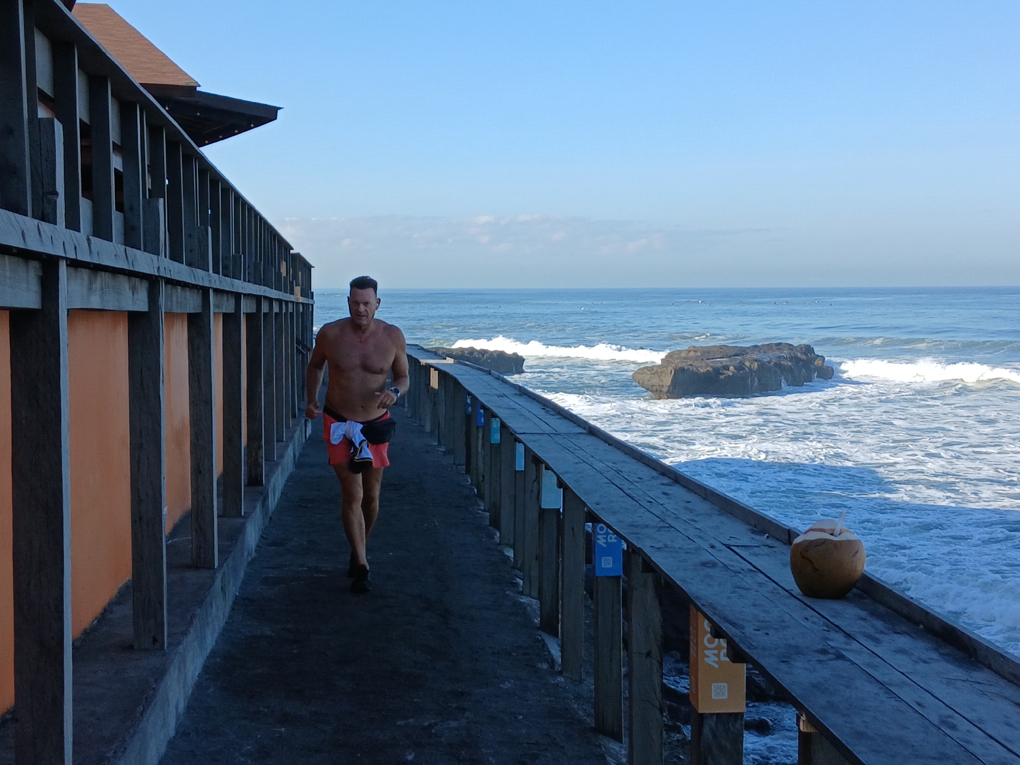 A shirtless man with short dark hair, wearing red and white swim shorts and a black fanny pack, is jogging along a wooden boardwalk beside the ocean. The boardwalk has a wooden railing and a building with an orange wall is visible to the left. The ocean is a vibrant blue with white waves crashing against rocks. The sky is clear and blue, with a few distant clouds. A coconut with a straw is placed on the boardwalk to the right. The scene appears to be a coastal path in North Kuta, Indonesia.