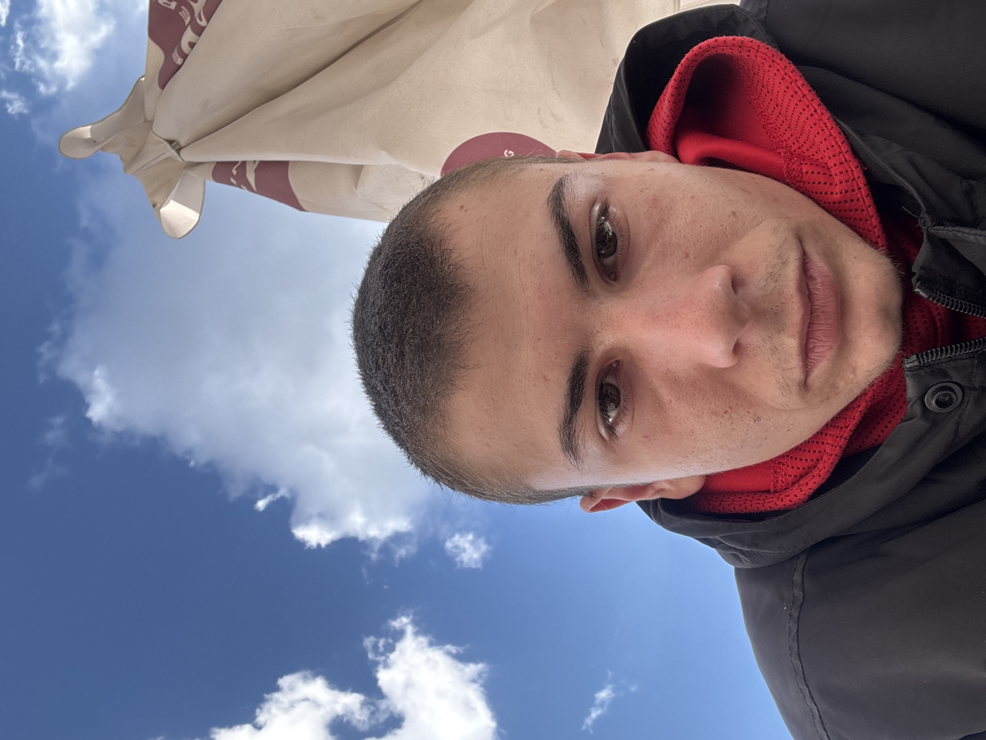 The image is a selfie of a man with short hair, brown eyes, wearing a black jacket with a red hoodie underneath. The background is a blue sky with white clouds and part of a tan-colored umbrella. The location is potentially a café or outdoor area in Plovdiv, Bulgaria, based on the typical umbrella designs found in outdoor dining spaces. There are no actions visible other than the subject taking a selfie.