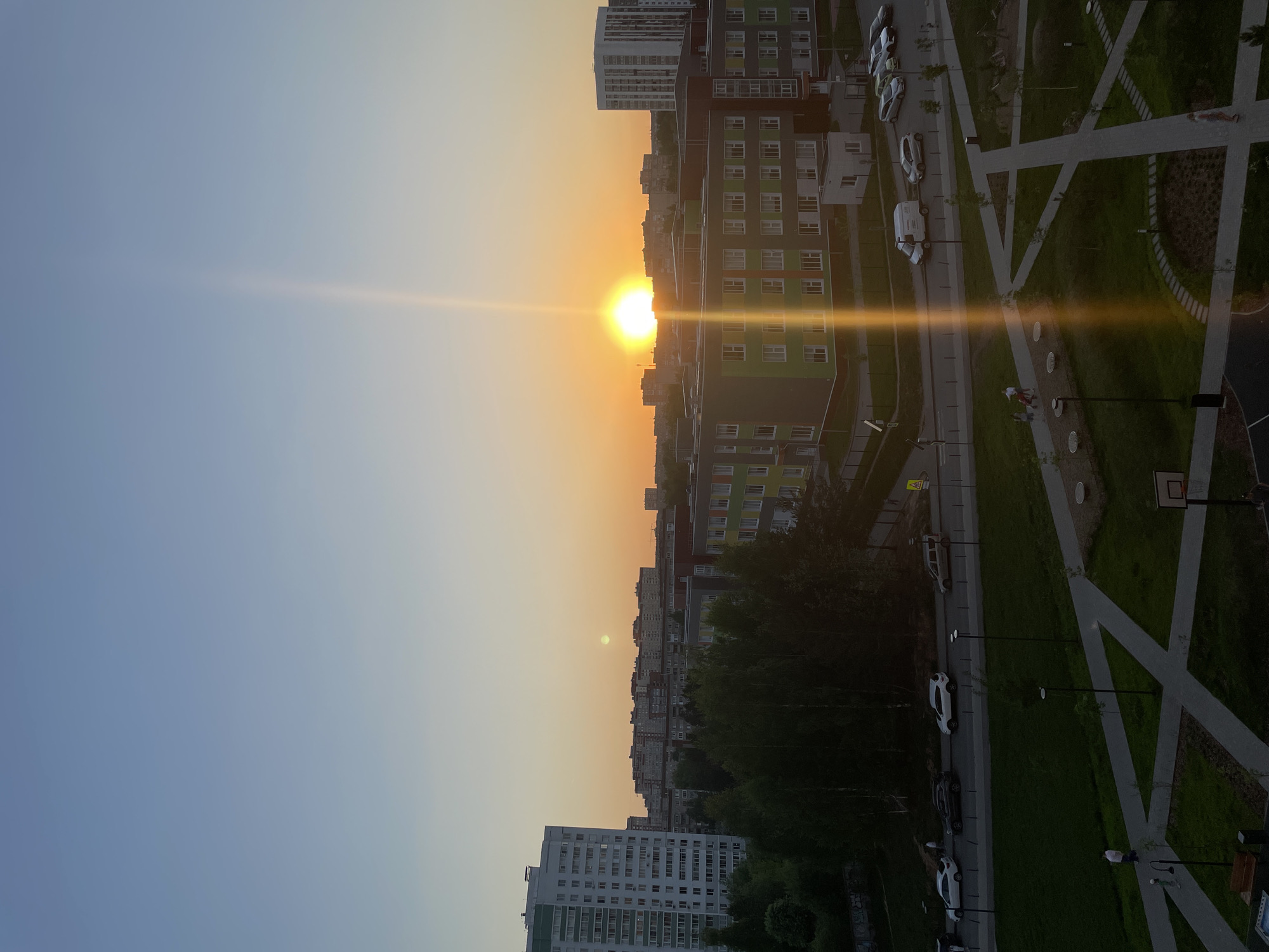 The image depicts a sunset over a cityscape, likely in Izhevsk, Russia. The sun is centered, causing a strong lens flare. Tall apartment buildings are visible on either side, with a park including walkways, street lights, and a basketball hoop on the right. Several parked cars are also present. The sky is blue in the upper left, transitioning to orange near the sun.