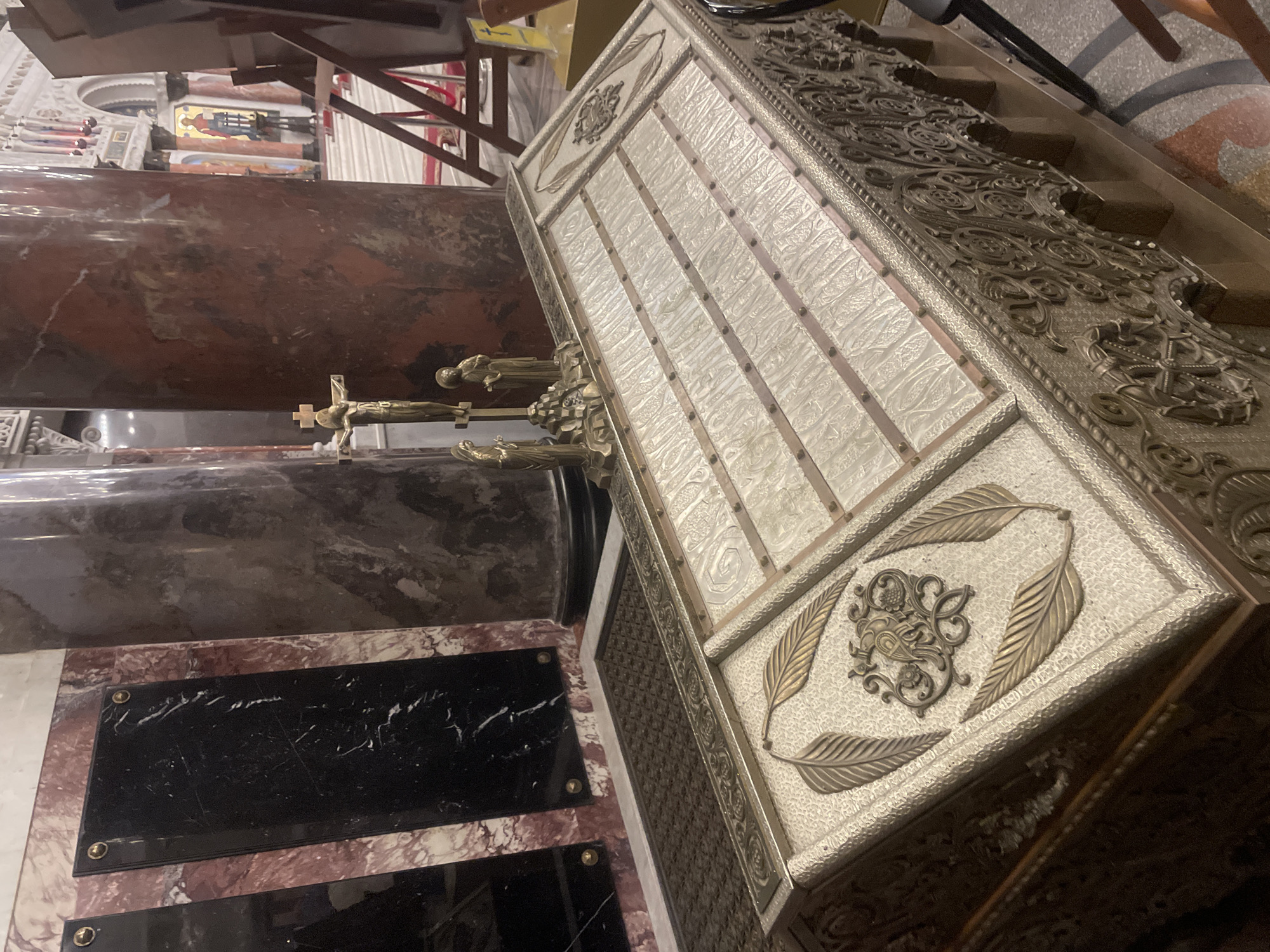 An ornate sarcophagus, possibly containing the remains of a notable figure, is the central focus of this image, situated within the St. Nicholas Naval Cathedral in Kronstadt, Russia. The sarcophagus is constructed from a metallic material, likely bronze or brass, and is adorned with intricate carvings and decorative elements. Its lid is segmented, with each section featuring a raised, textured pattern and metal bands. The front and sides of the sarcophagus are elaborately decorated with repeating arched motifs and ornate scrollwork.
Adjacent to the sarcophagus, a small crucifix with figurines of Jesus Christ, Mary, and John the Apostle stands, suggesting a religious context and possibly commemorating a saintly or reverent individual. In the background, large marble columns with reddish-brown veining rise, typical of the cathedral's architecture. Dark marble plaques are visible on the wall to the left, hinting at memorial inscriptions. Further in the background, the interior of the cathedral is partially visible, with architectural details, religious iconography, and a red carpet runner indicating a sacred space. The overall impression is one of solemnity and historical significance.