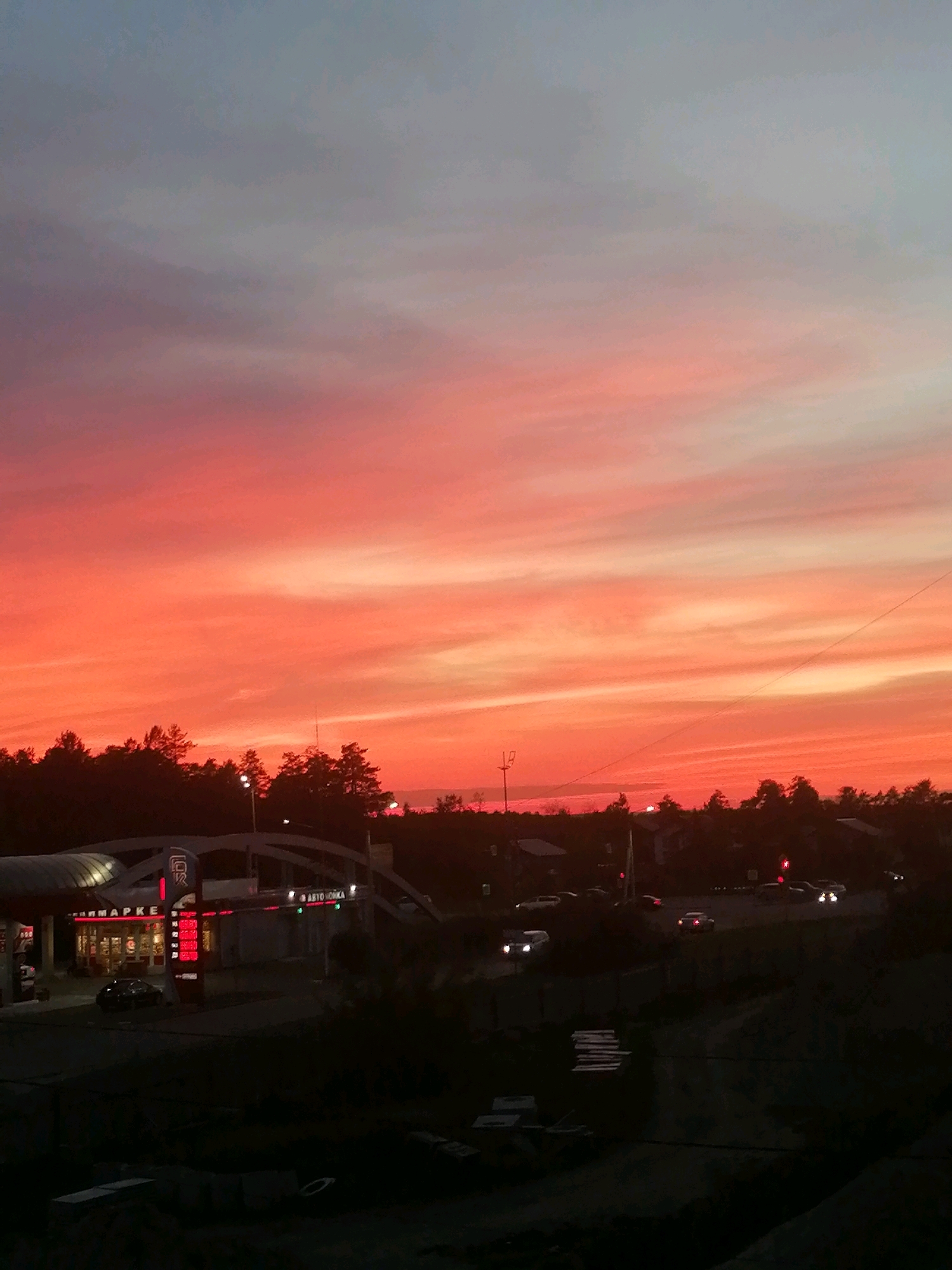 The image captures a vibrant sunset over Angarsk, Russia. The sky is a dramatic gradient of colors, transitioning from a pale blue at the top to intense shades of orange, pink, and red near the horizon. Wispy clouds are illuminated by the setting sun, creating streaks of light across the sky.
In the foreground, silhouetted against the bright sky, is a landscape featuring a line of trees. Below the trees, a gas station with illuminated signage is visible on the left side of the image. The signs display red lettering, including the word "MARKET". Several cars are parked at the gas station and on the road in front of it, with their headlights on. Further to the right, more cars are driving on what appears to be a road or intersection, their headlights also illuminated. The overall impression is of a typical evening scene in a semi-urban or suburban environment, with the natural beauty of the sunset contrasting with the man-made structures and activity.