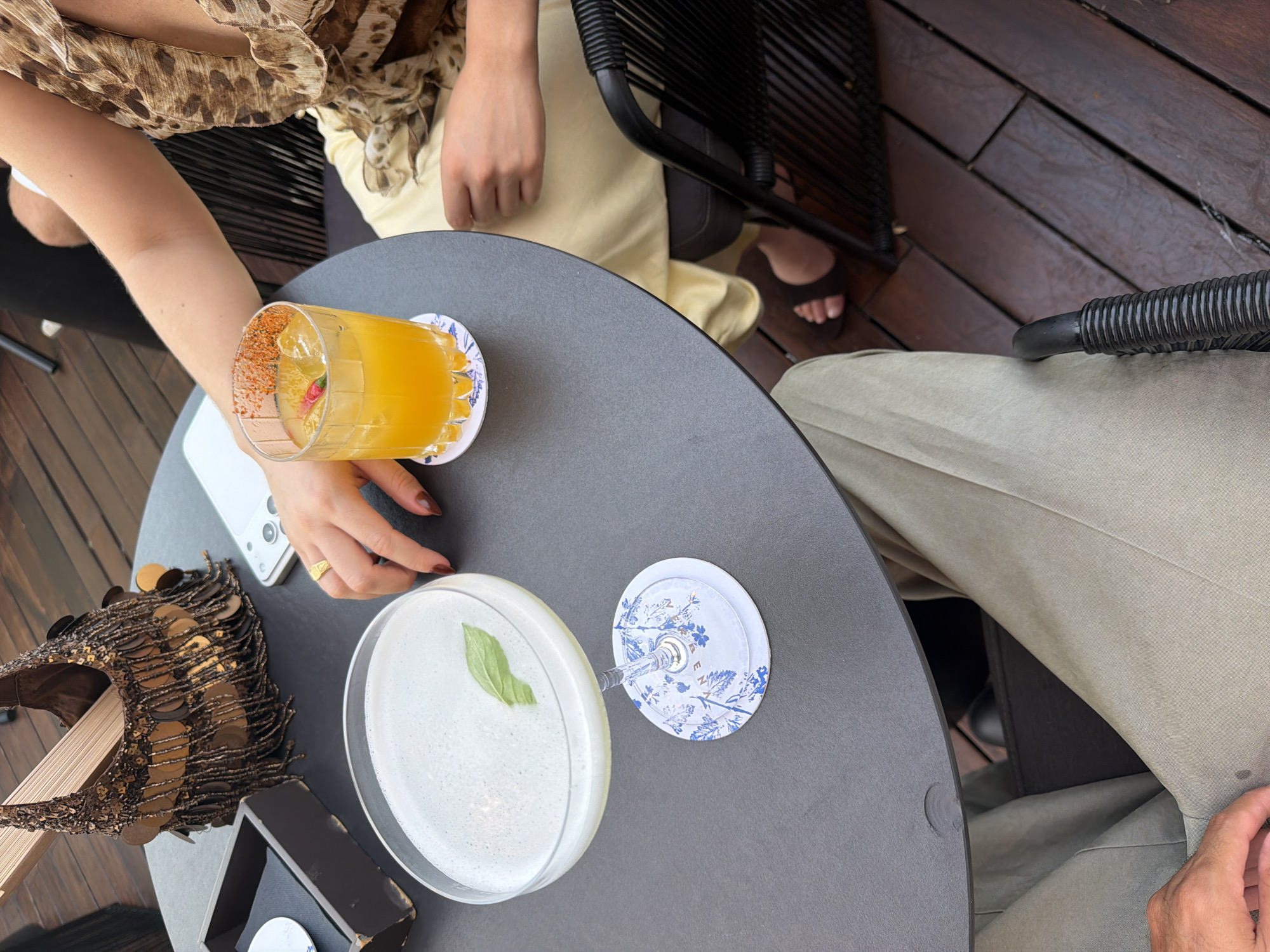 At a table outdoors, two individuals are seated with drinks and a handbag. One person, wearing a leopard-print top and a yellow skirt, holds a cocktail with ice and a garnish of red pepper and a pink flower. Their hand rests on the table near the drink. The other individual, wearing khaki pants, has their hand resting on their lap. A second cocktail, a pale frothy drink with a mint leaf, sits on a decorative coaster on the table, next to a small black box and a metallic handbag with fringe. A white smartphone is also visible on the table. The scene is set on a wooden deck, suggesting an outdoor dining or lounge area. The image lacks specific details to confirm a location within Barcelona, Spain.