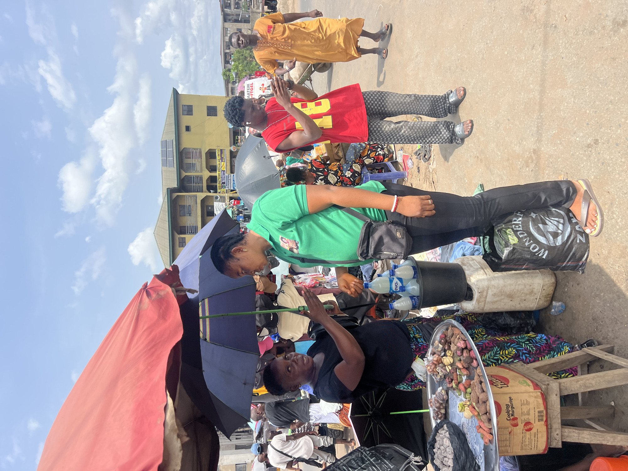 The image depicts a bustling street scene in Onitsha, Nigeria, likely a marketplace area. A woman in a black shirt stands behind a stall, shaded by an umbrella, selling what appears to be spices and root vegetables displayed on a tray. A carton of "Cooking Margarine" is visible beside the tray. A woman in a green shirt stands next to her, seemingly observing or interacting. Behind them, a crowd of people navigates the busy street. Two men are standing on the side of the street in a red and yellow shirt respectively. The background shows buildings, including one with solar panels, against a partly cloudy sky. The environment is dusty, with various objects scattered around, including water bottles and plastic containers.