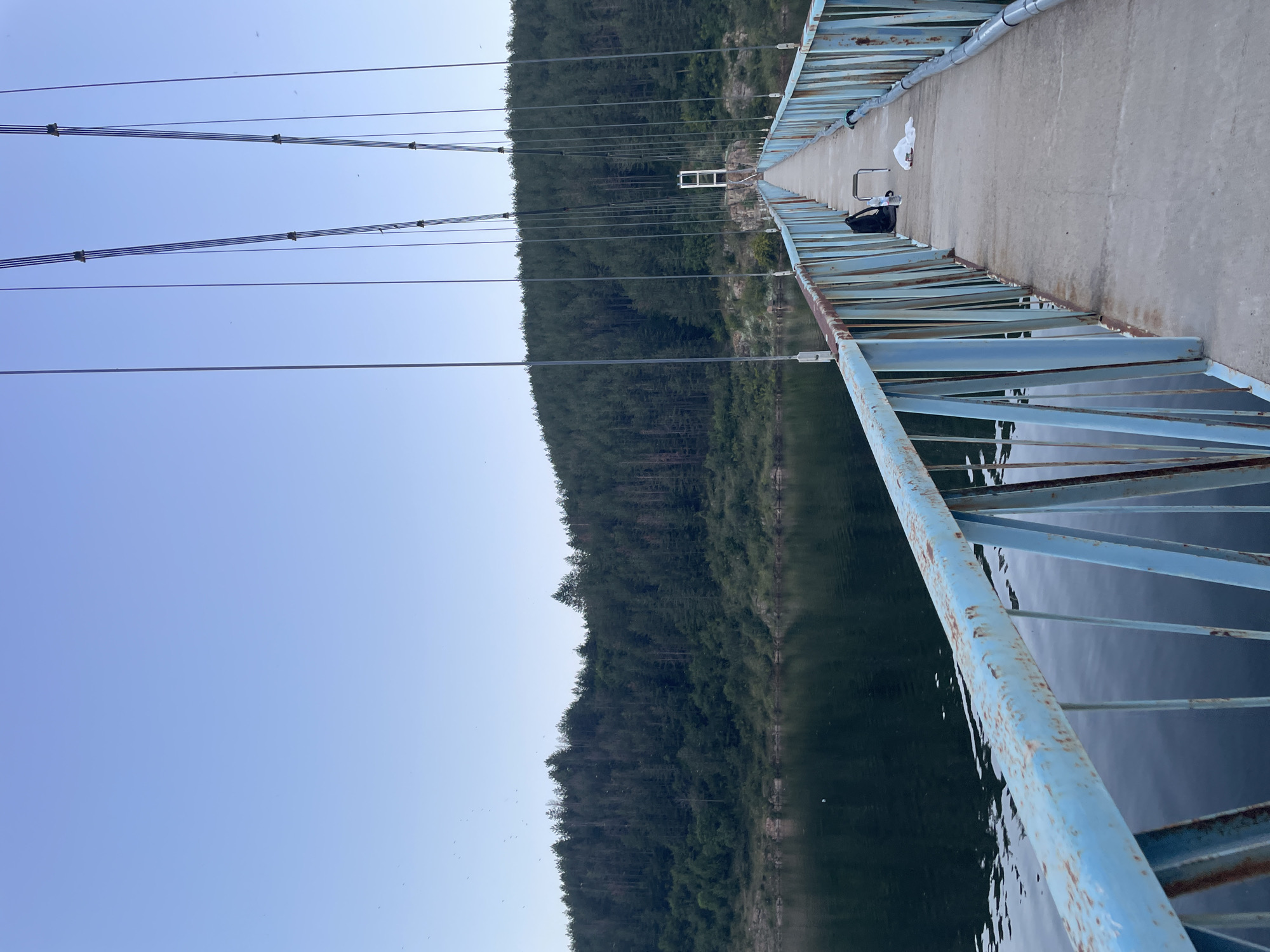 A perspective shot from a light blue metal-railed pedestrian suspension bridge shows a concrete walkway extending into the distance towards a forest-covered mountain range. The bridge's blue railings have visible rust spots. On the walkway, a black backpack and a white bag are placed near a small folding chair. To the left of the walkway, a dark body of water reflects the muted colors of the sky and the trees. The sky is a clear, pale blue. The forest on the mountain is dense and green, with some trees appearing bare. The overall scene suggests a serene, natural setting. The bridge is likely located in Suhovo, Bulgaria, connecting to the area around the dam.
