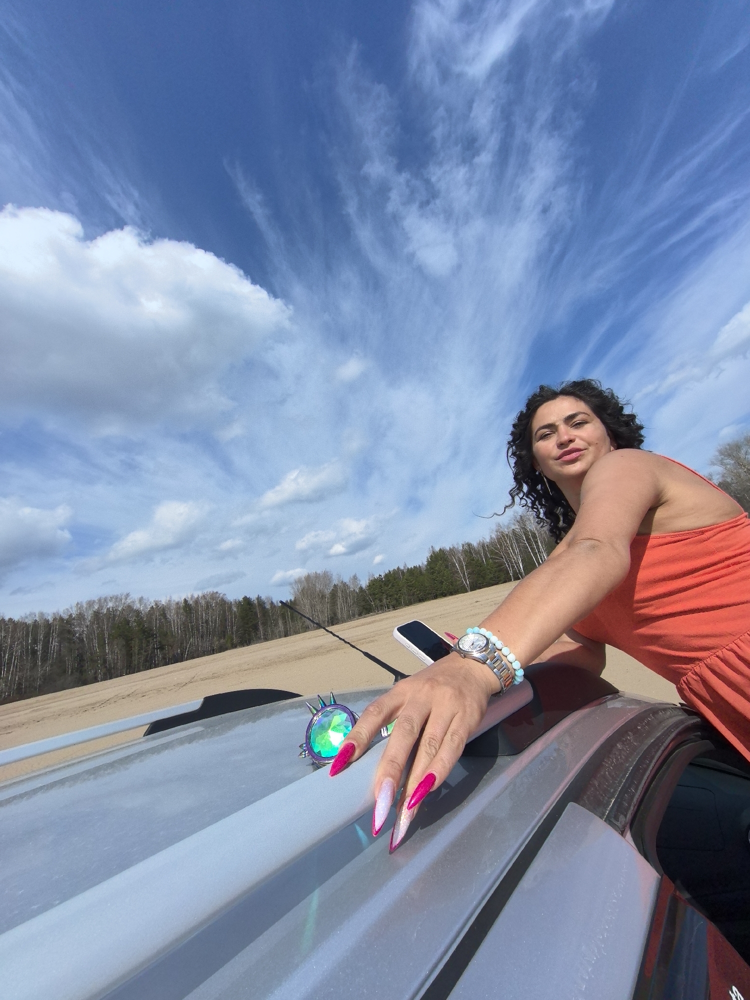 A woman with curly dark hair and an orange sundress leans on the roof of a silver car, looking directly at the camera with a slight smile. Her right arm is extended across the car's roof. She has long, pointed fingernails, painted in a vibrant pink with a white or light pink gradient. On her left wrist, she wears a silver watch and two beaded bracelets, one light blue and one white/beige. A smartphone rests on the car roof near her left hand, which is also adorned with the distinctive manicure. Beside her hand, on the car roof, there is a small, iridescent, faceted decorative object, possibly a car accessory. A car antenna is visible extending from the roof towards the right side of the frame.
The setting is outdoors, likely a sunny daytime scene in Chaikovsky, Russia. The background features a large expanse of light brown, sandy ground, resembling a beach or dune. Behind the sandy area, a dense line of trees, including both leafless deciduous trees and evergreen conifers, forms a forest. The sky is a prominent feature, displaying a bright blue hue with scattered white clouds, ranging from large, puffy cumulus clouds to delicate, wispy cirrus clouds stretching across the frame. No discernible text is visible in the image.