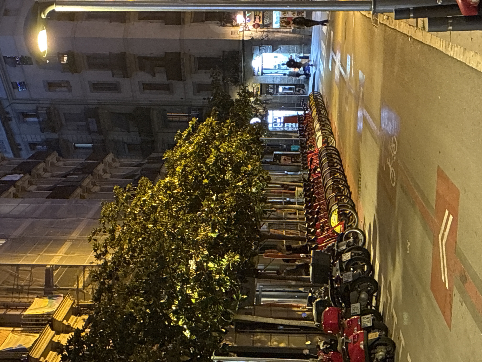 This nocturnal image captures an urban street scene in Barcelona, Spain. The street, paved with asphalt, stretches into the background, illuminated by overhead streetlights and the glow from distant shopfronts.
Along the right side of the street, a long array of red and black rental bicycles or scooters is neatly parked, forming a prominent sharing station. Several vehicles display legible license plates, including "9382 JPF," "9218 HL," and "1362 HTM." The street surface features painted markings, with a clear white arrow and a partial bicycle symbol indicating a dedicated lane.
To the left, a dense line of trees with vibrant green foliage runs parallel to the street, brightly lit by yellow streetlights which highlight their leaves. Beyond the trees and extending along both sides of the street, multi-story buildings rise, showcasing architectural details like windows and balconies. Some structures, particularly on the far left, appear to be under renovation, with scaffolding visible.
In the distance, a few blurry figures of people can be seen near brightly lit shopfronts, suggesting commercial activity. One storefront clearly displays an "ATM" sign. An overhead streetlight positioned towards the top left of the frame casts a strong light down the street. The overall ambiance is one of a quiet, functional city street at night, with various modes of transport readily available.